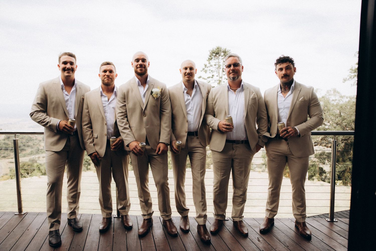 Groomsmen in matching beige suits pose on wooden deck with scenic view.