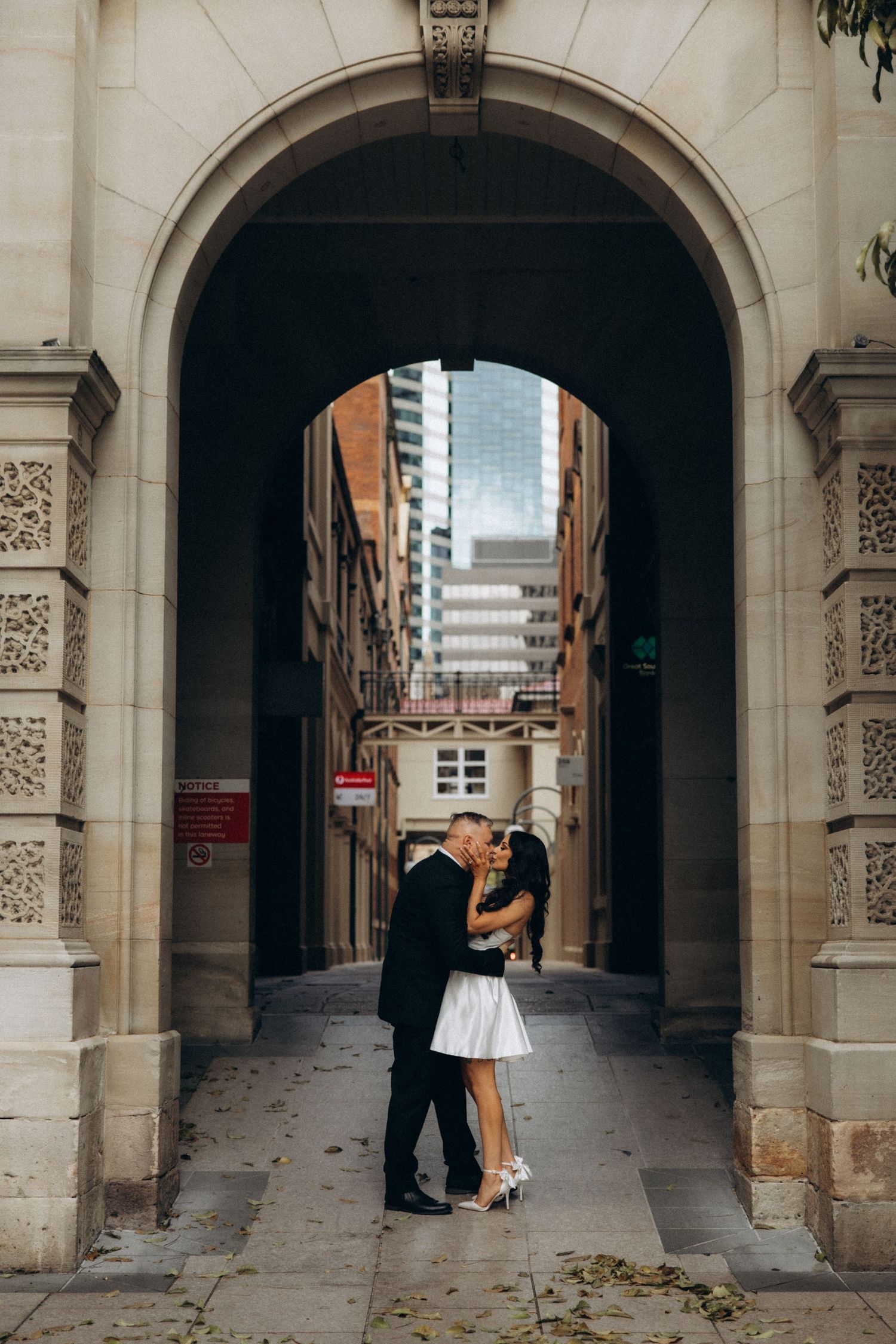 Couple sharing a romantic kiss under a grand stone archway in an urban setting, with city buildings visible behind them.