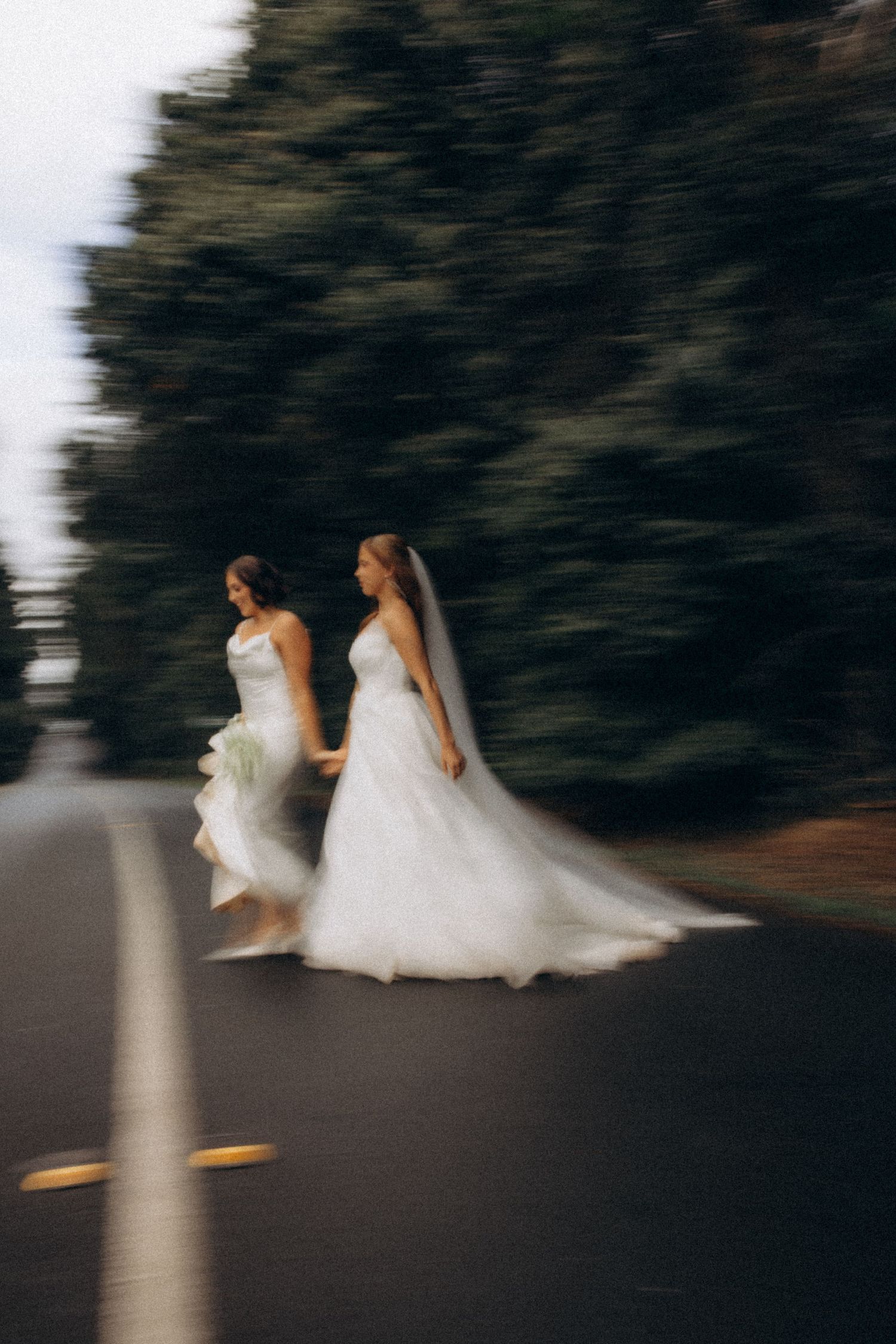 Motion-blurred photograph of two brides walking in white wedding dresses on a dark road.