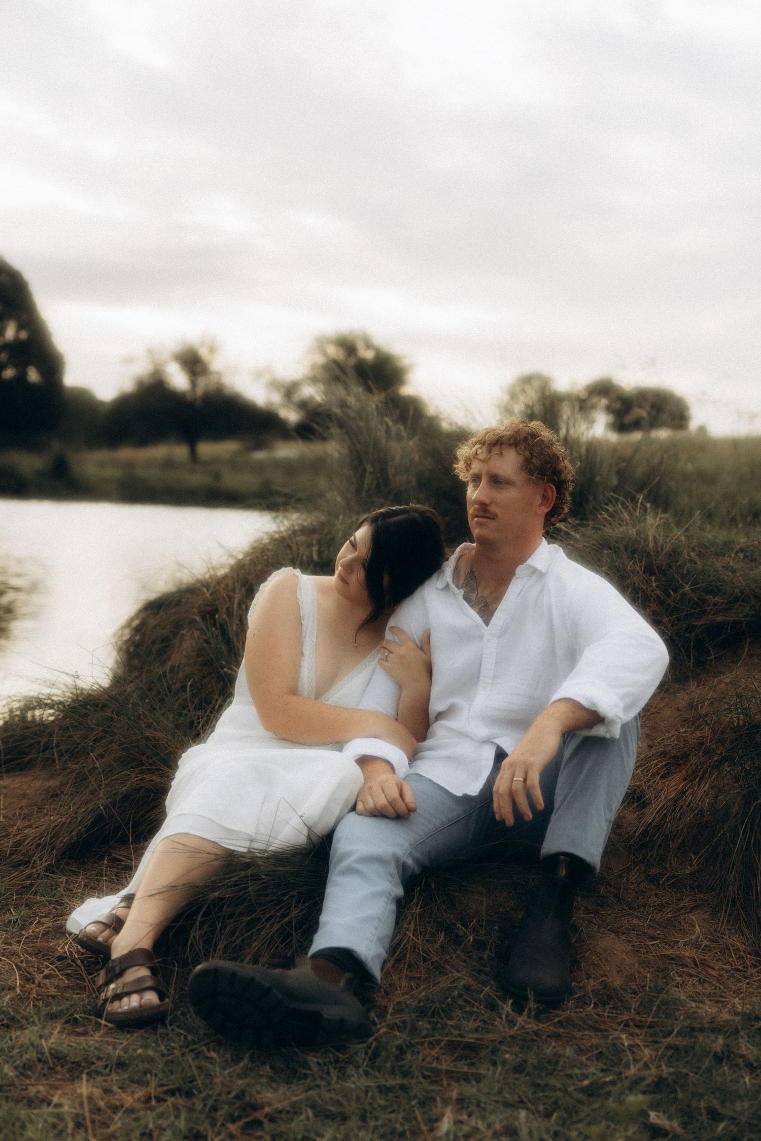 Couple in white clothing lounging together on a hillside near water.