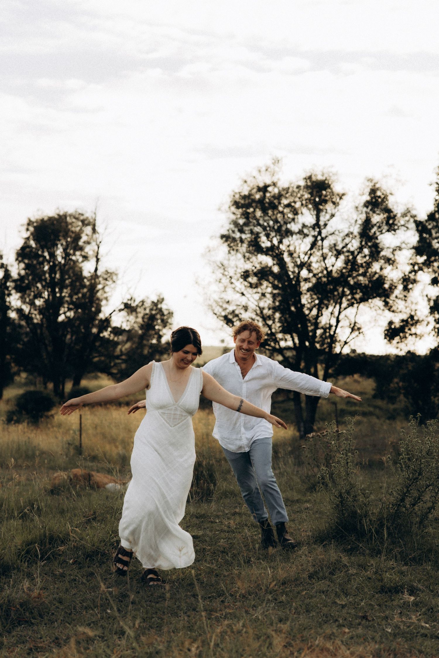 A couple dances playfully in a field at sunset with trees in the background.