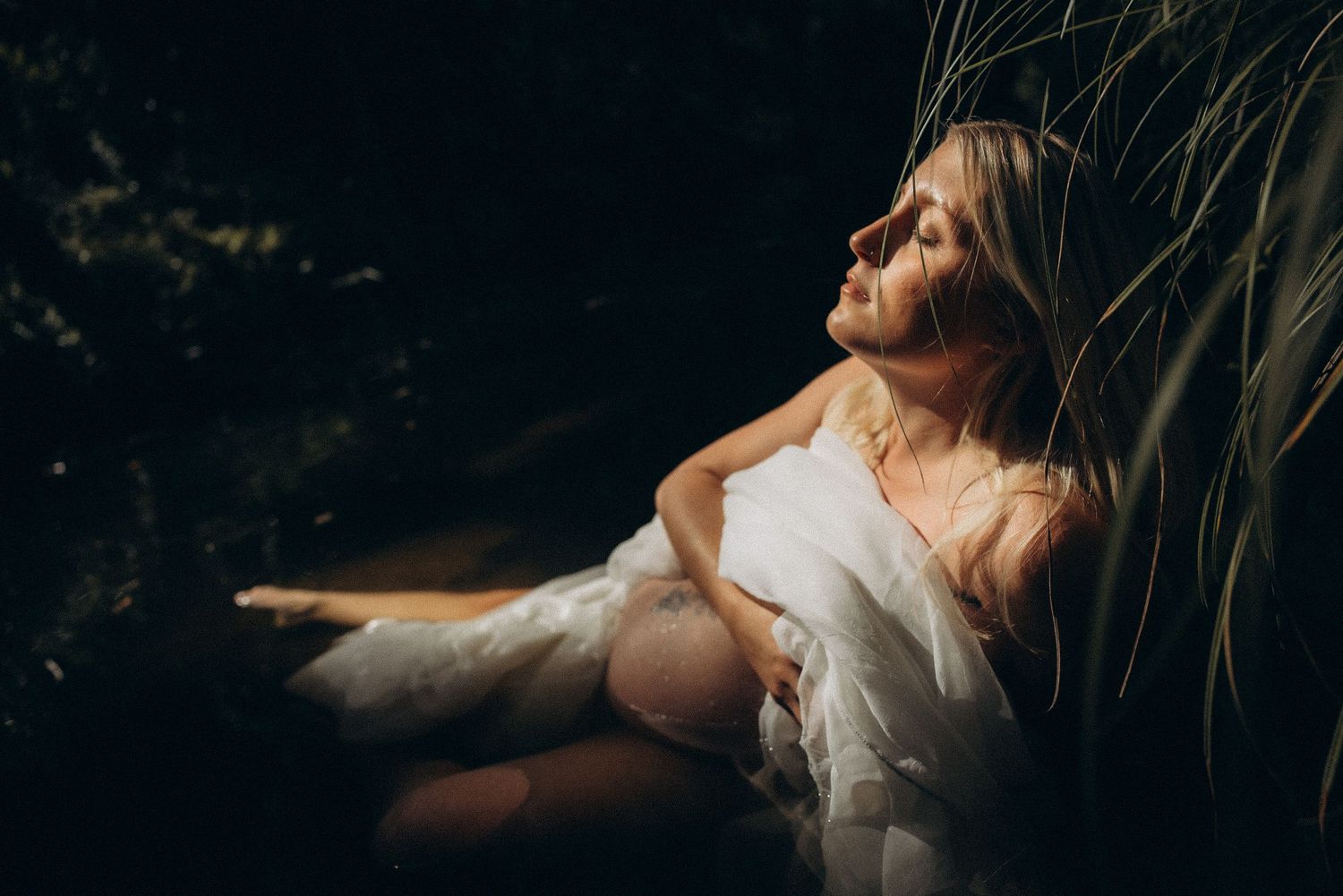 Pregnant woman in white dress relaxing outdoors against lush greenery in soft natural light.
