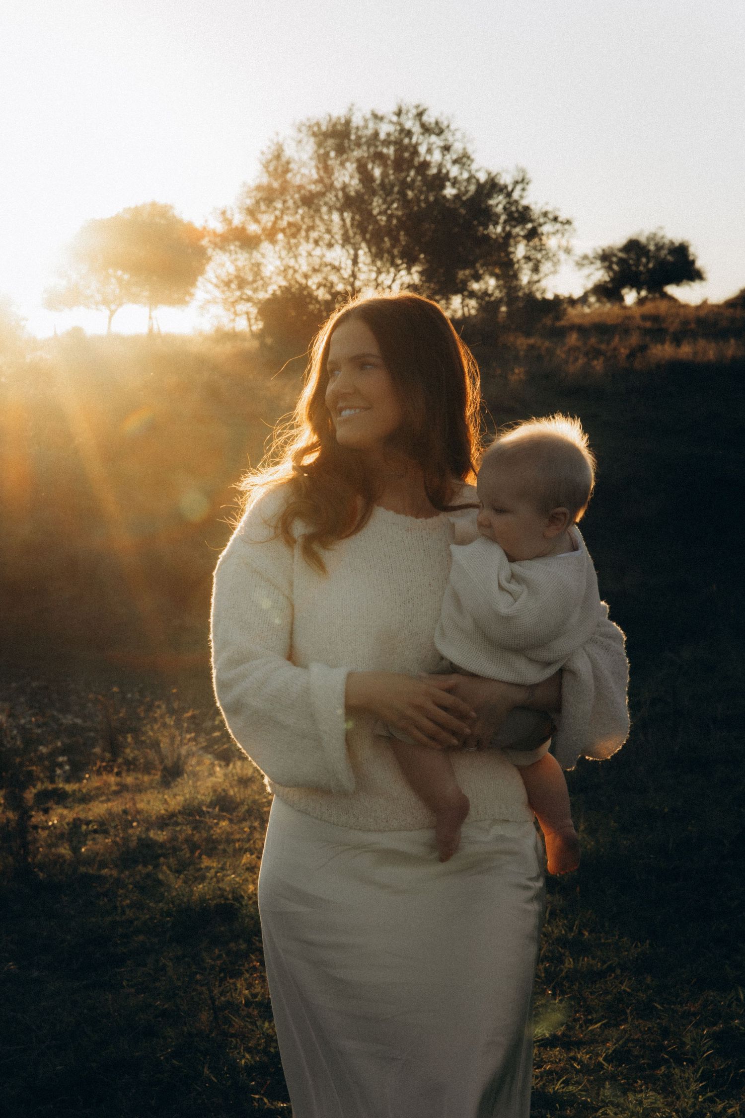 A smiling woman in white holds a baby outdoors during golden hour sunset with warm light and trees behind.