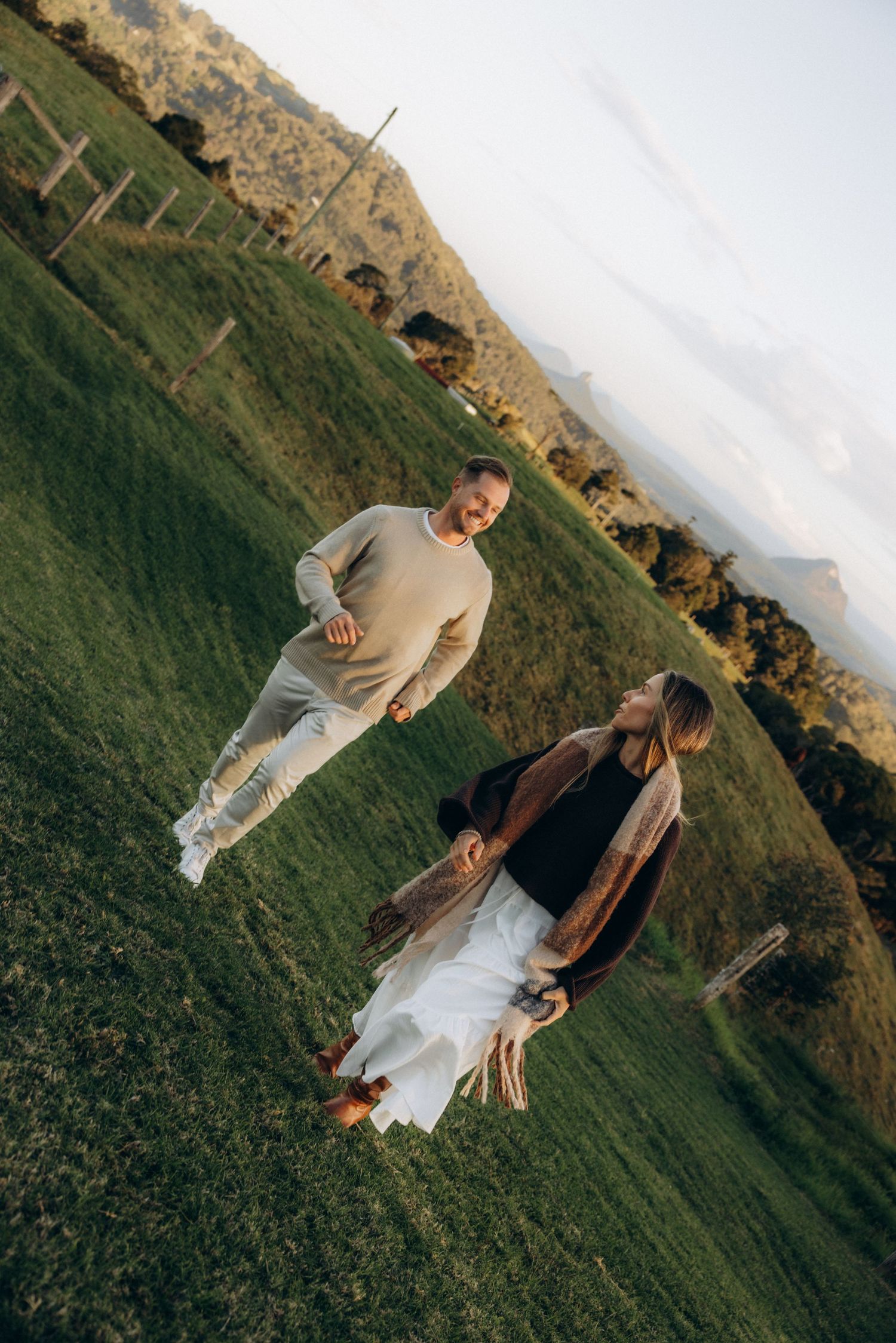 Couple walking hand in hand on lush green hillside at golden hour, smiling and enjoying a romantic outdoor moment.