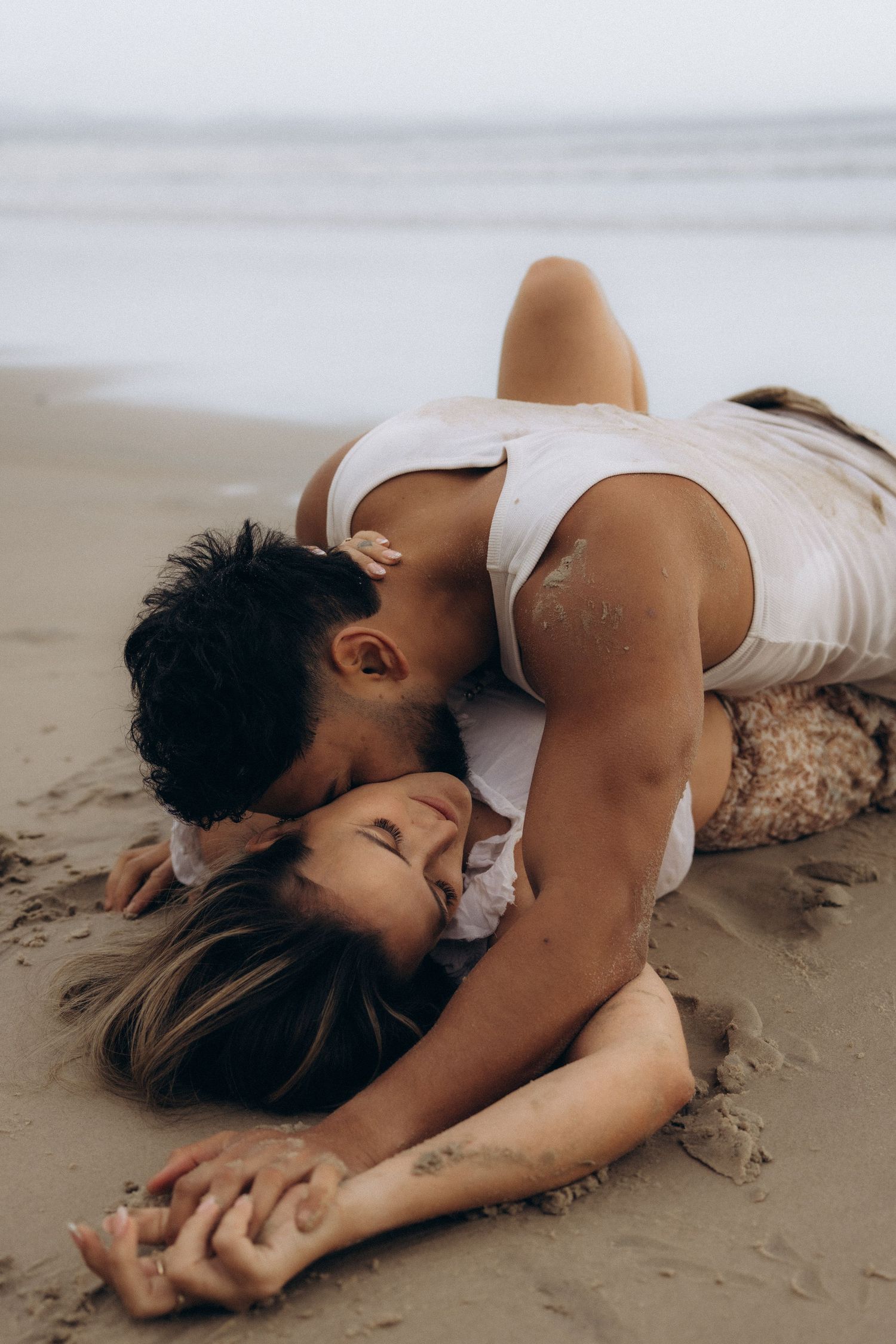 Couple sharing a romantic kiss on a sandy beach, lying together in an intimate embrace by the ocean shoreline.
