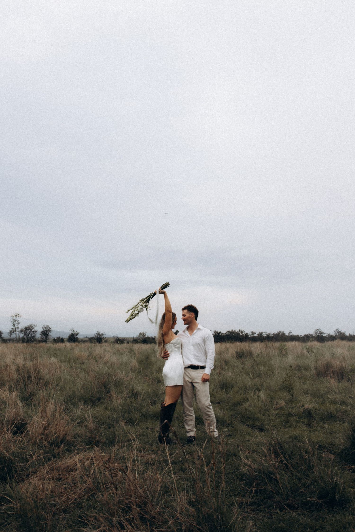 Wedding couple celebrates joyfully in an open field under cloudy skies.