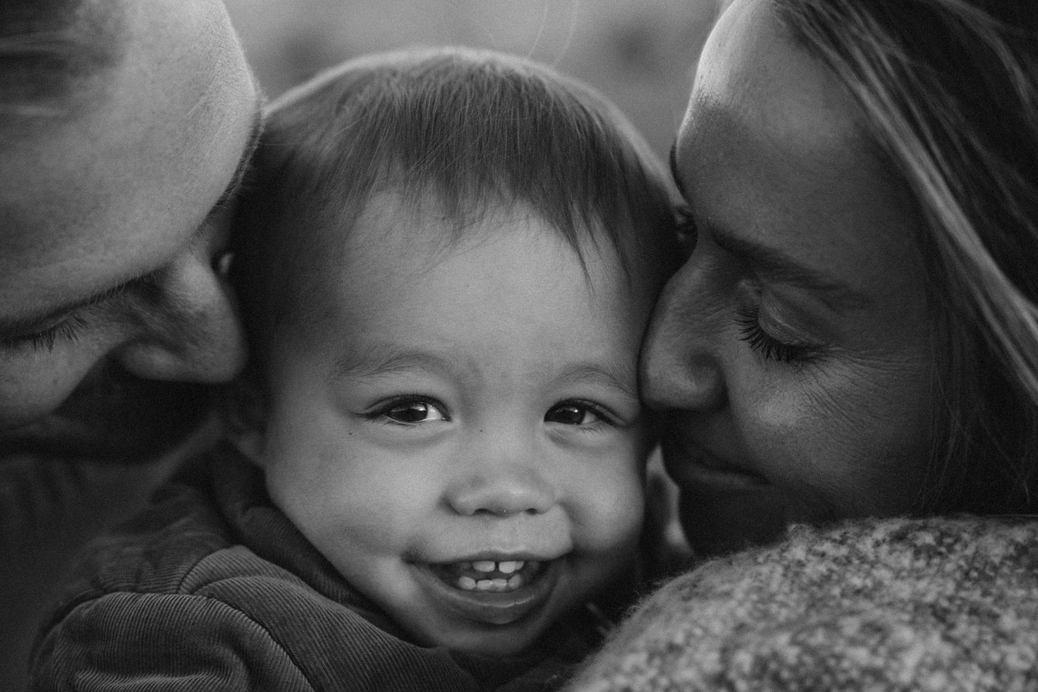 Black and white photo of smiling baby being kissed on cheeks by both parents in a tender family moment.