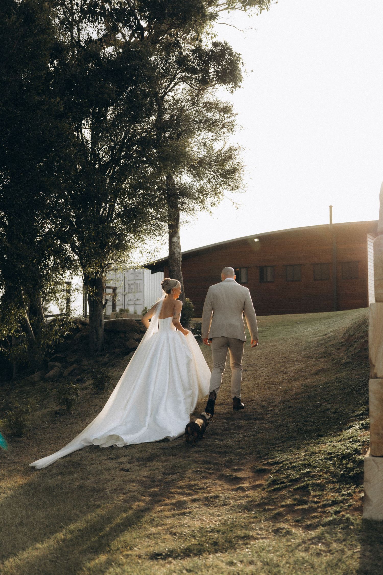 Couple walking up a grassy hill at sunset with bride's dress trailing behind.