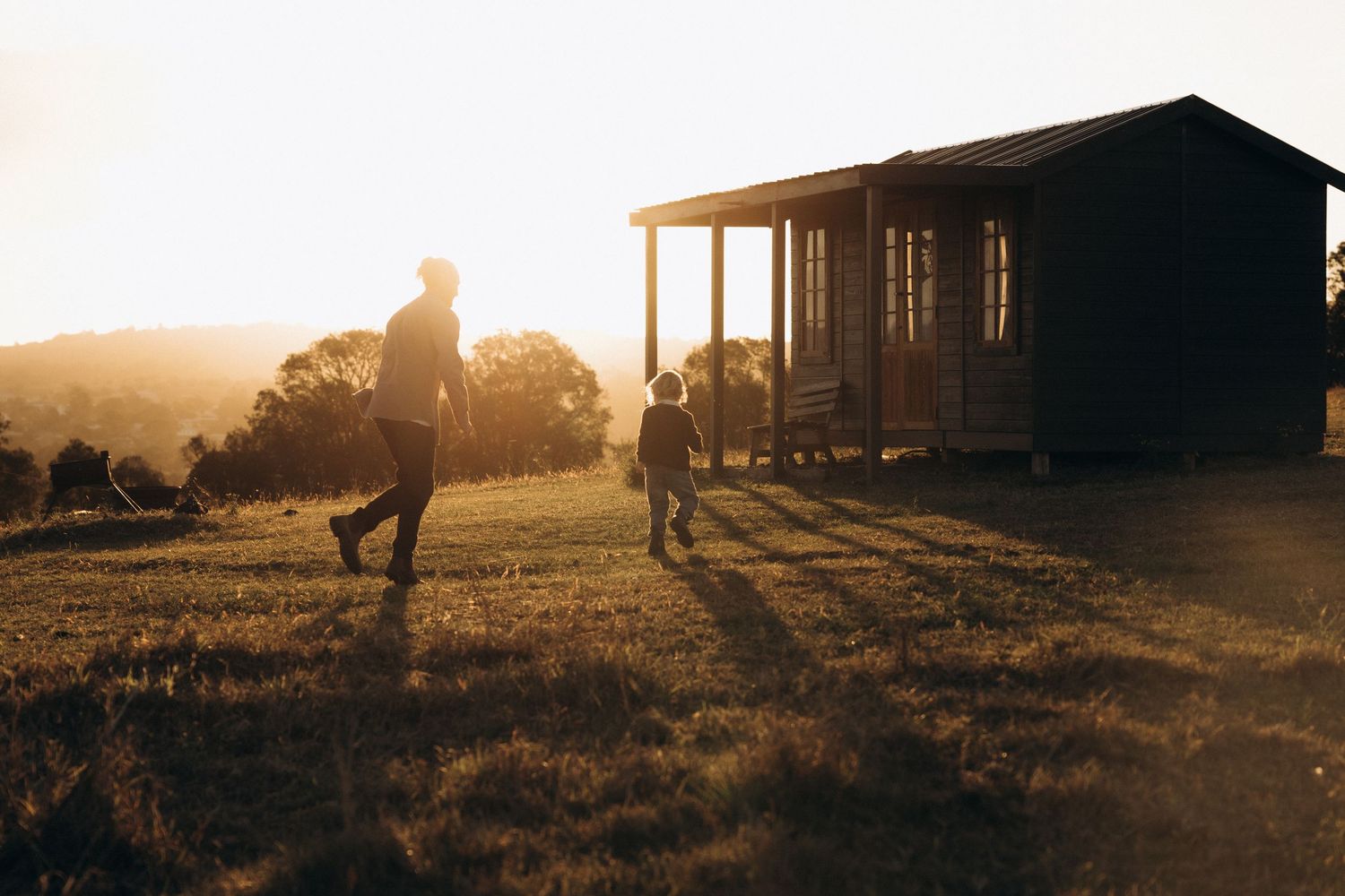 Two silhouettes walk toward a rustic cabin at golden sunset on a peaceful rural property.