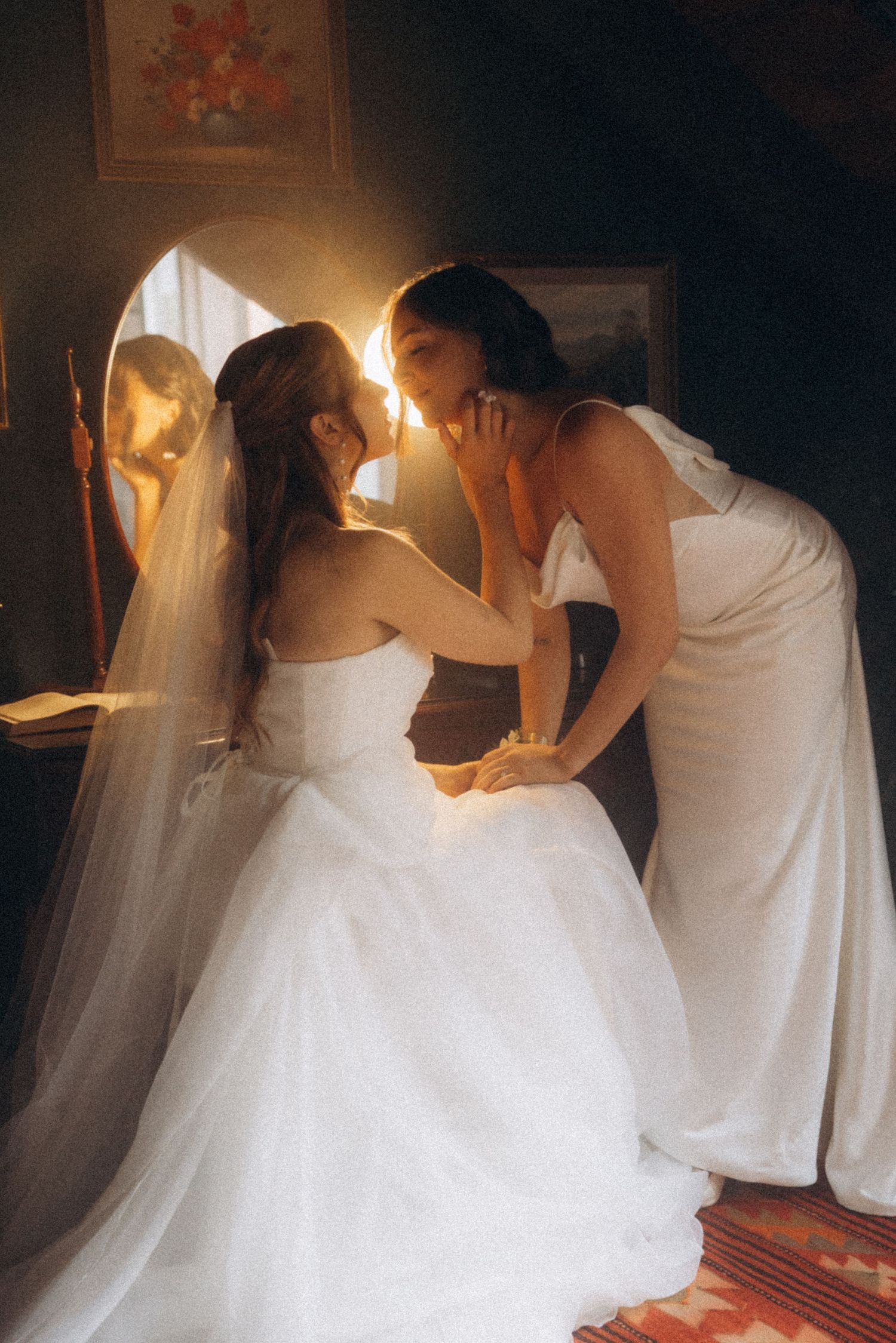 Two brides in white gowns share an intimate moment by a mirror in warm golden light.