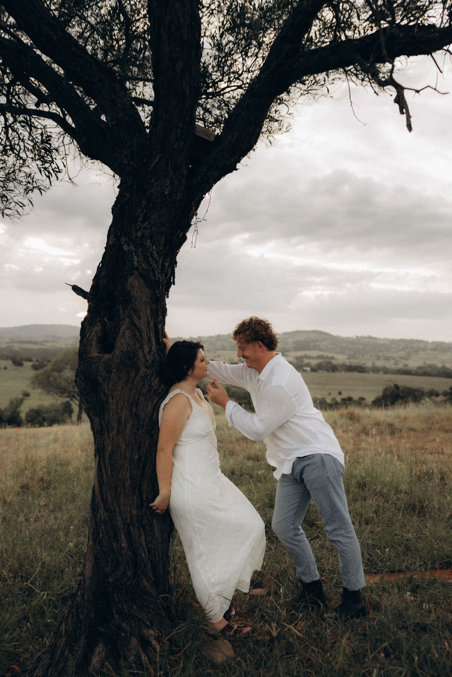 Two people share playful interactions around an old tree in a grassy field.