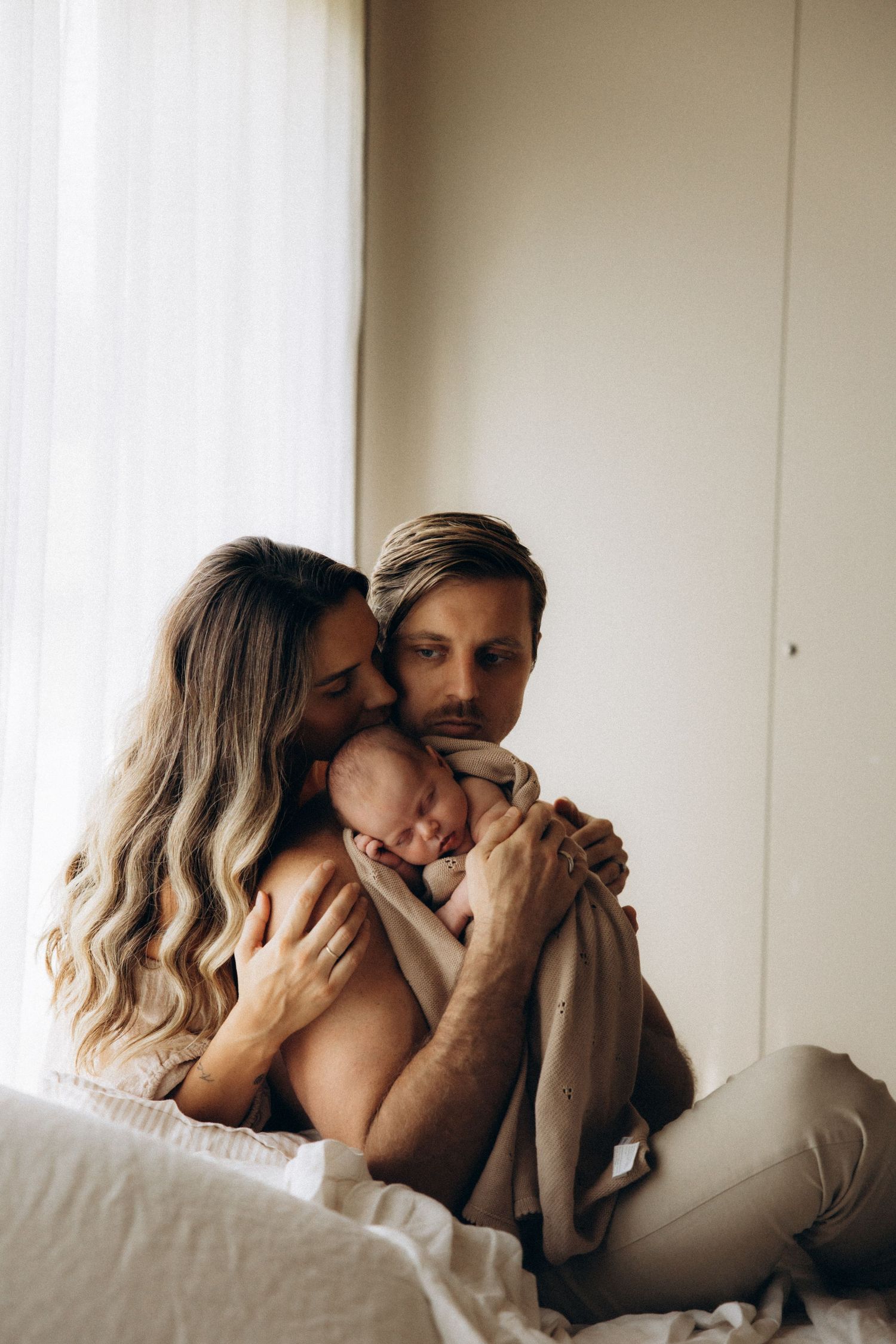Tender family moment as parents embrace sleeping newborn near bright window.