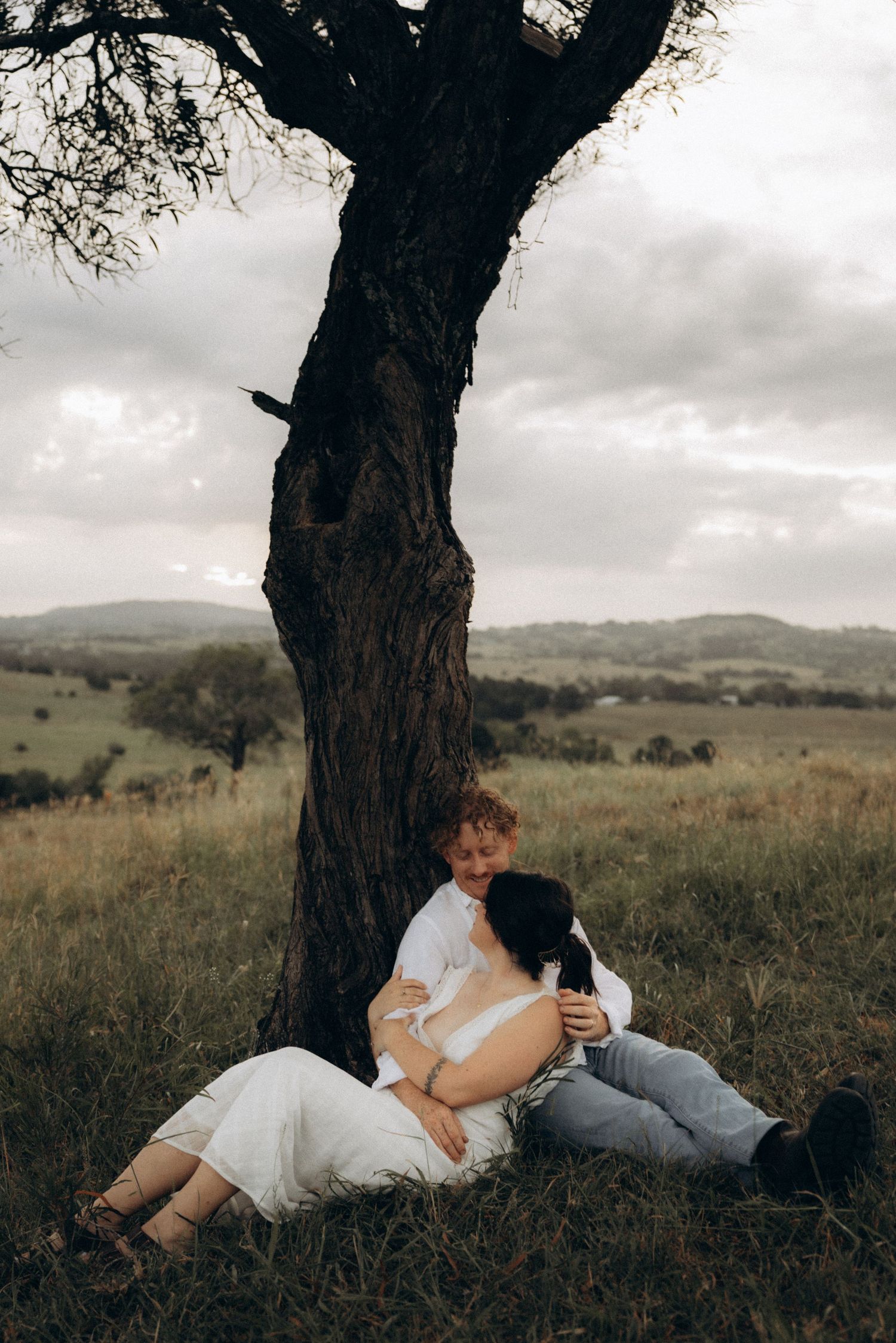 A couple reclines against a large tree trunk in a peaceful meadow setting.