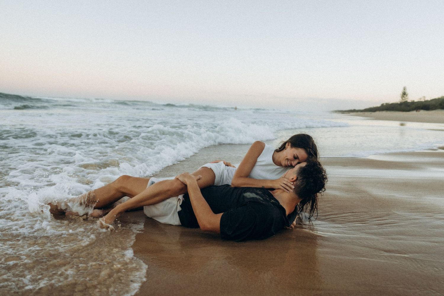 Romantic couple lying together in ocean waves on a sandy beach at sunset.