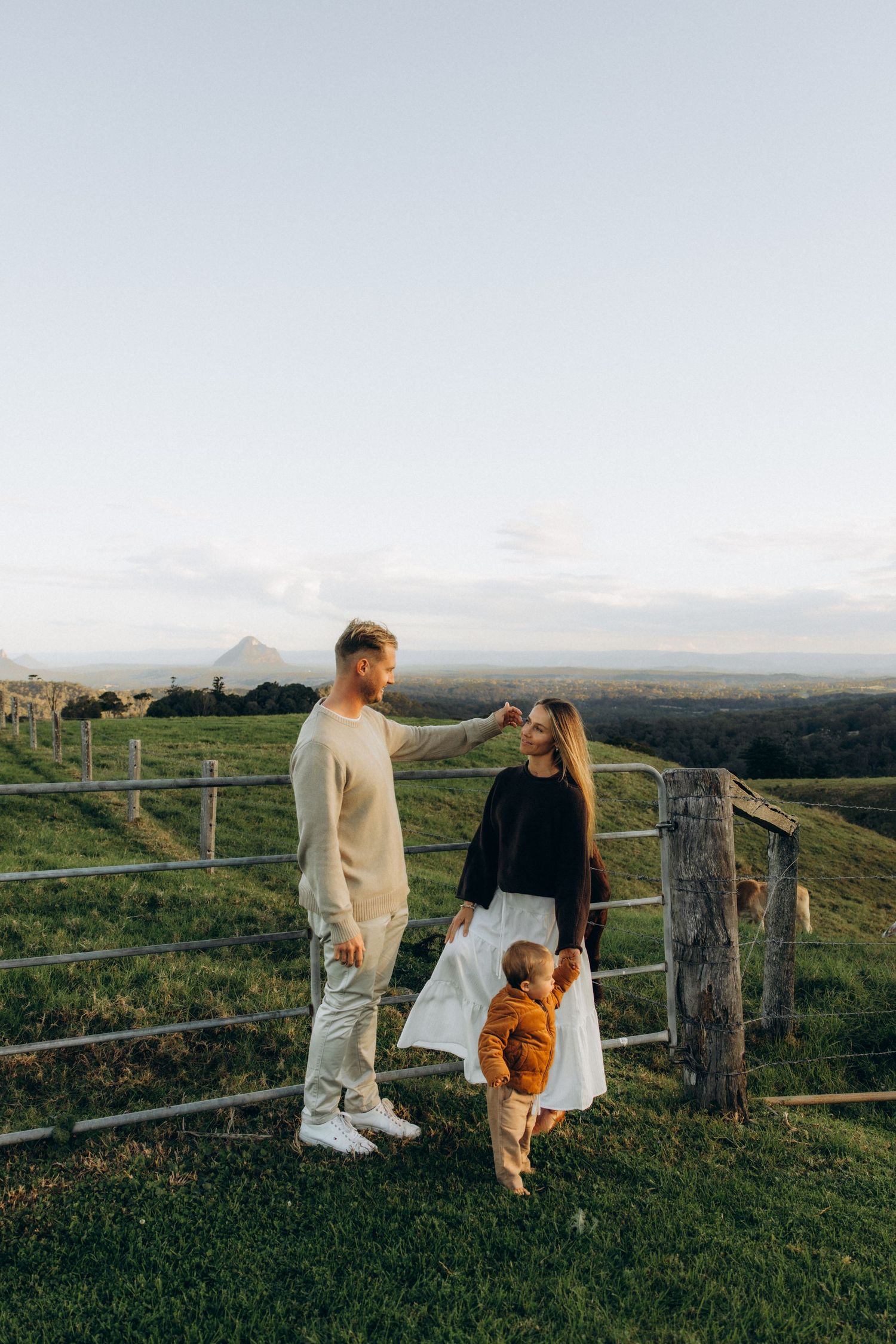 Family of three standing by a farm gate at golden hour, with green fields and a mountain in the background.