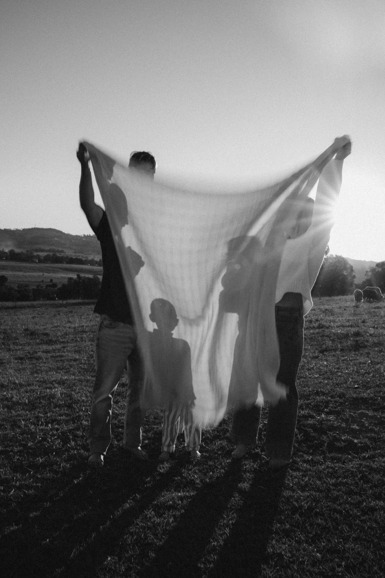 Black and white photo of three people holding a sheer blanket backlit by sunset in an open field.