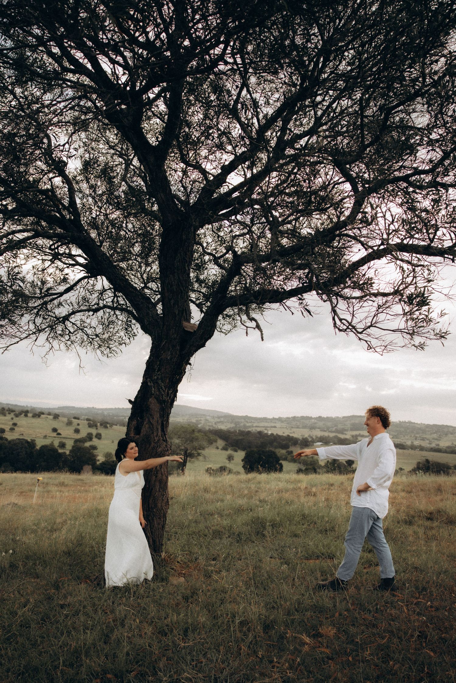 A couple shares a romantic moment beneath a sprawling tree in an open field.