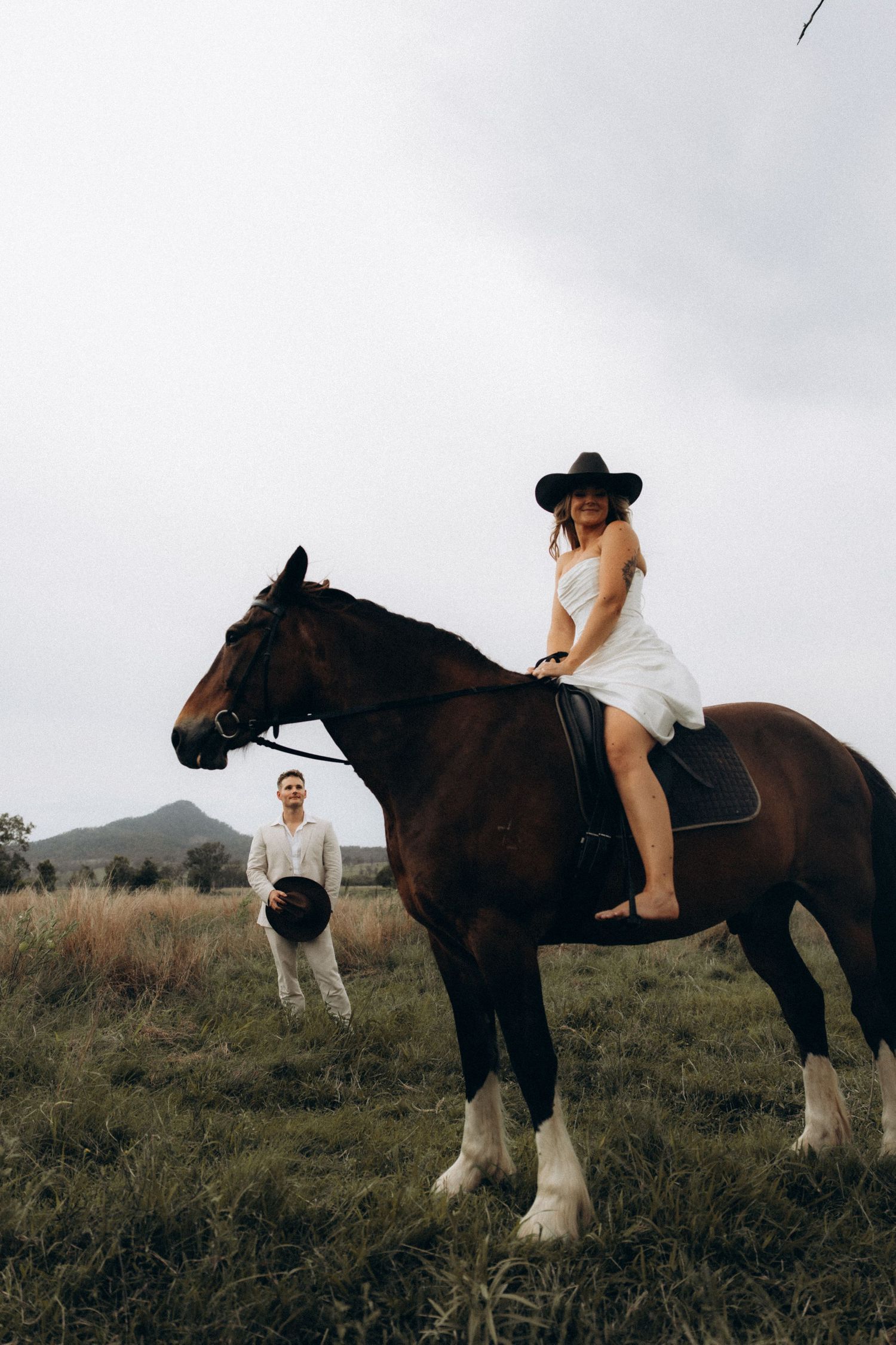 A person in a white dress rides a large draft horse while another stands nearby in a field.