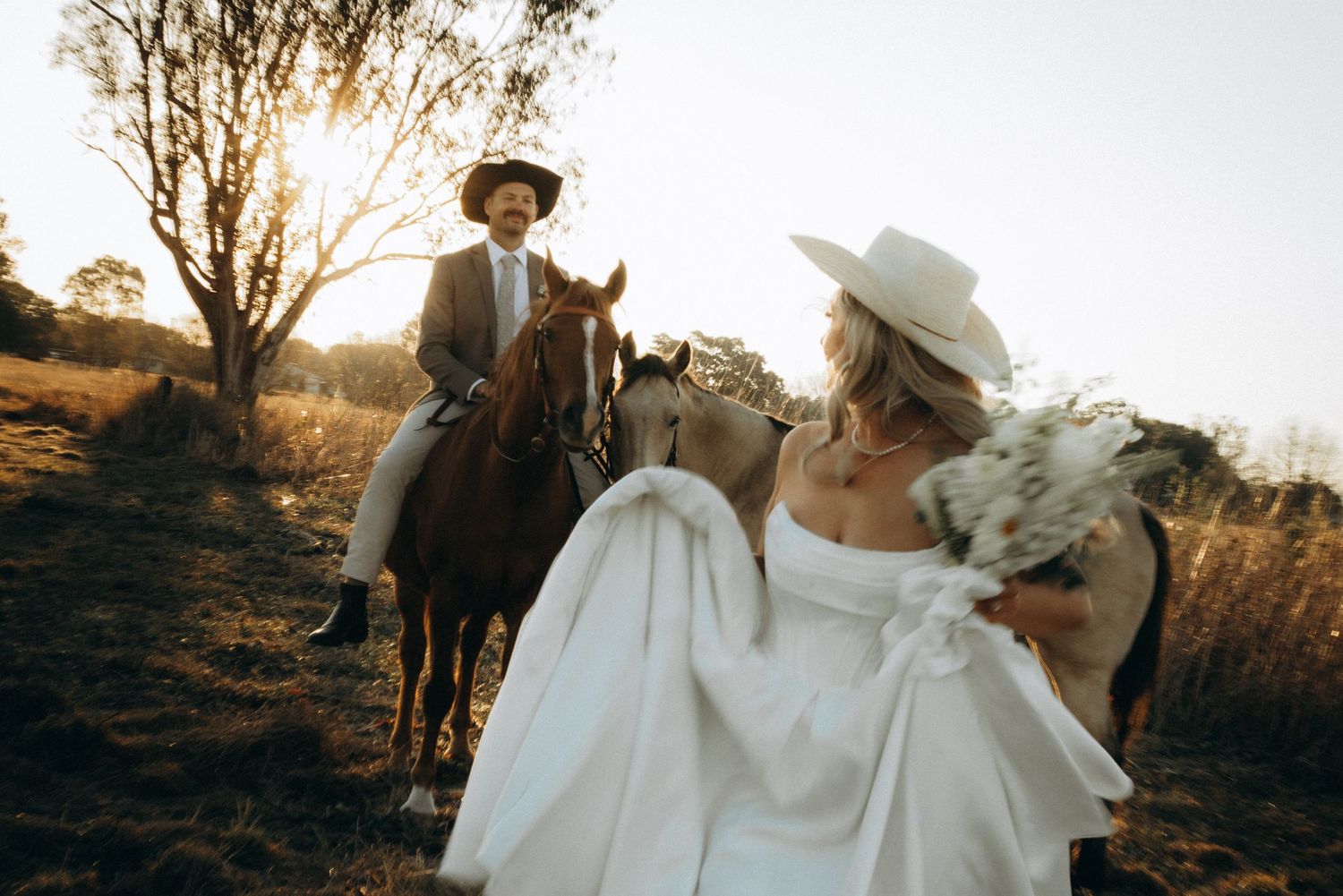 Bride in white gown and hat approaches groom on horseback at golden sunset during rustic outdoor wedding.