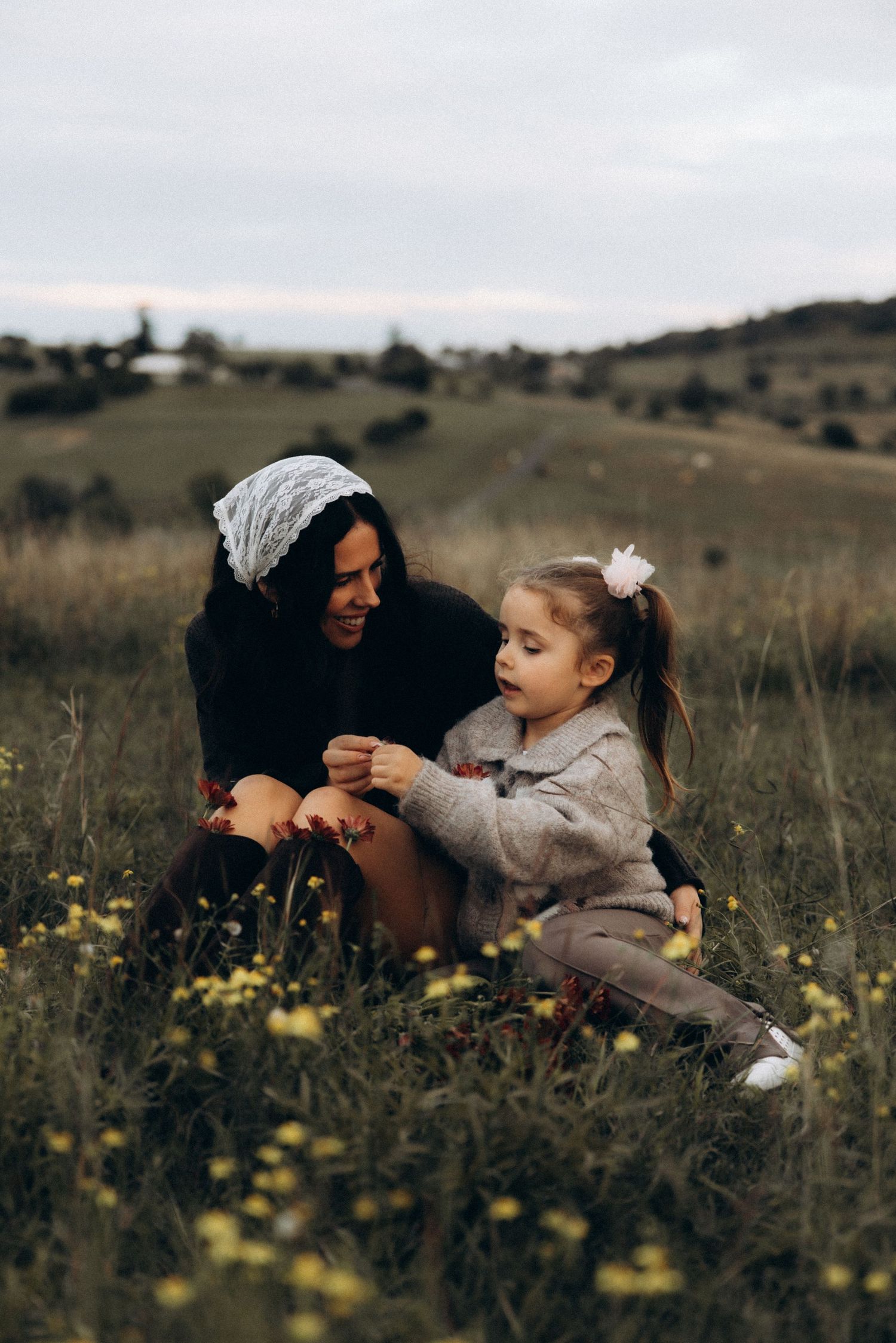 A mother and young daughter sit together in a field of yellow wildflowers, sharing a tender moment in a scenic countryside.