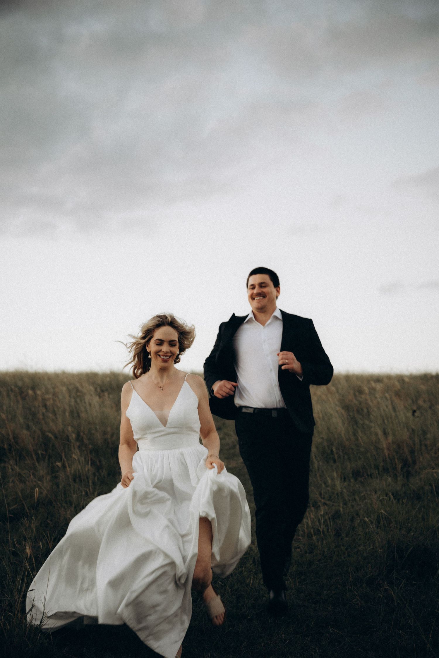 A joyful bride and groom run through a grassy field in wedding attire under a cloudy sky.