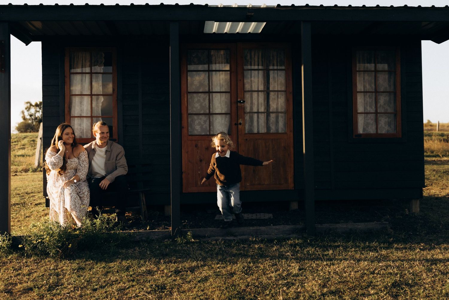 Family posing outside rustic dark wood cabin at golden hour, with child running on porch.