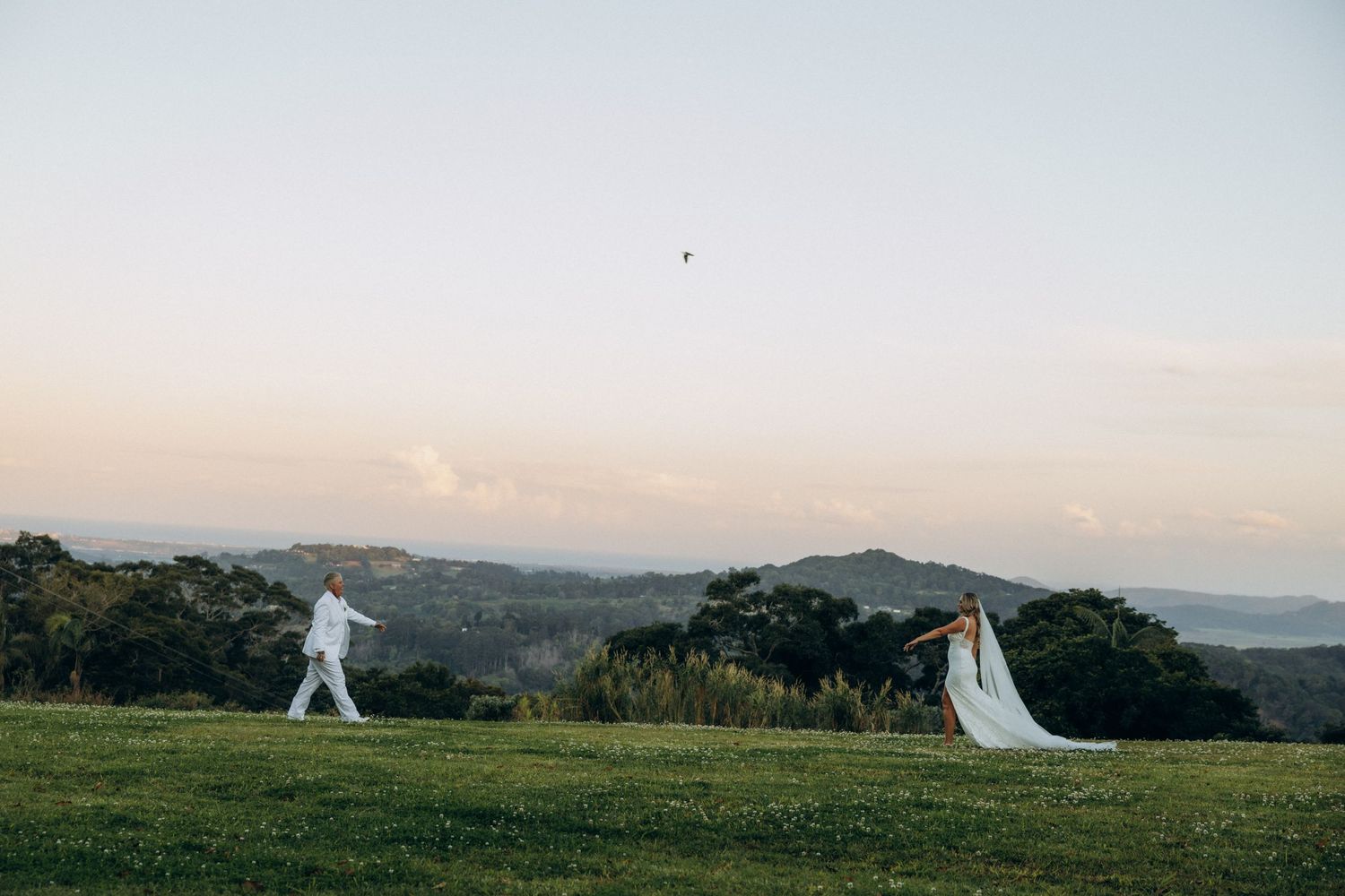 Bride and groom in white attire stand on a lush green hillside at sunset with scenic valley views.
