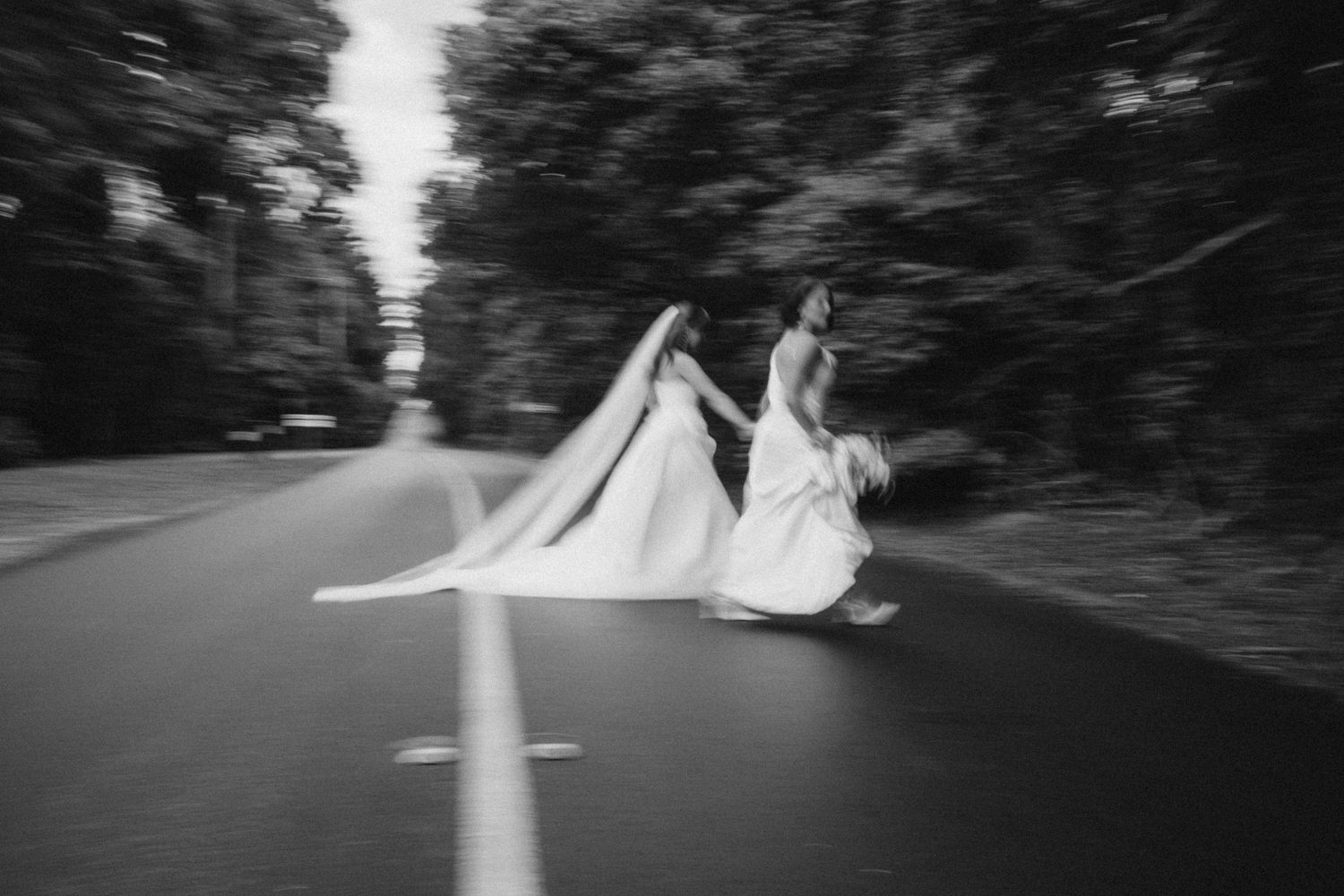 Two brides in white gowns run hand-in-hand down a tree-lined road in a blurred black and white wedding photo.