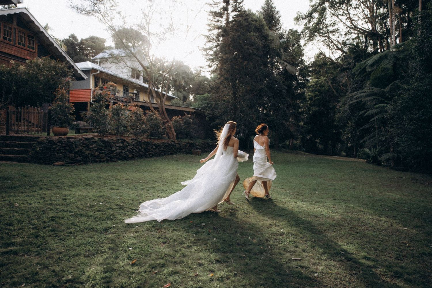 Bride in white gown walks with flower girl across lush green lawn near rustic venue surrounded by tall trees.