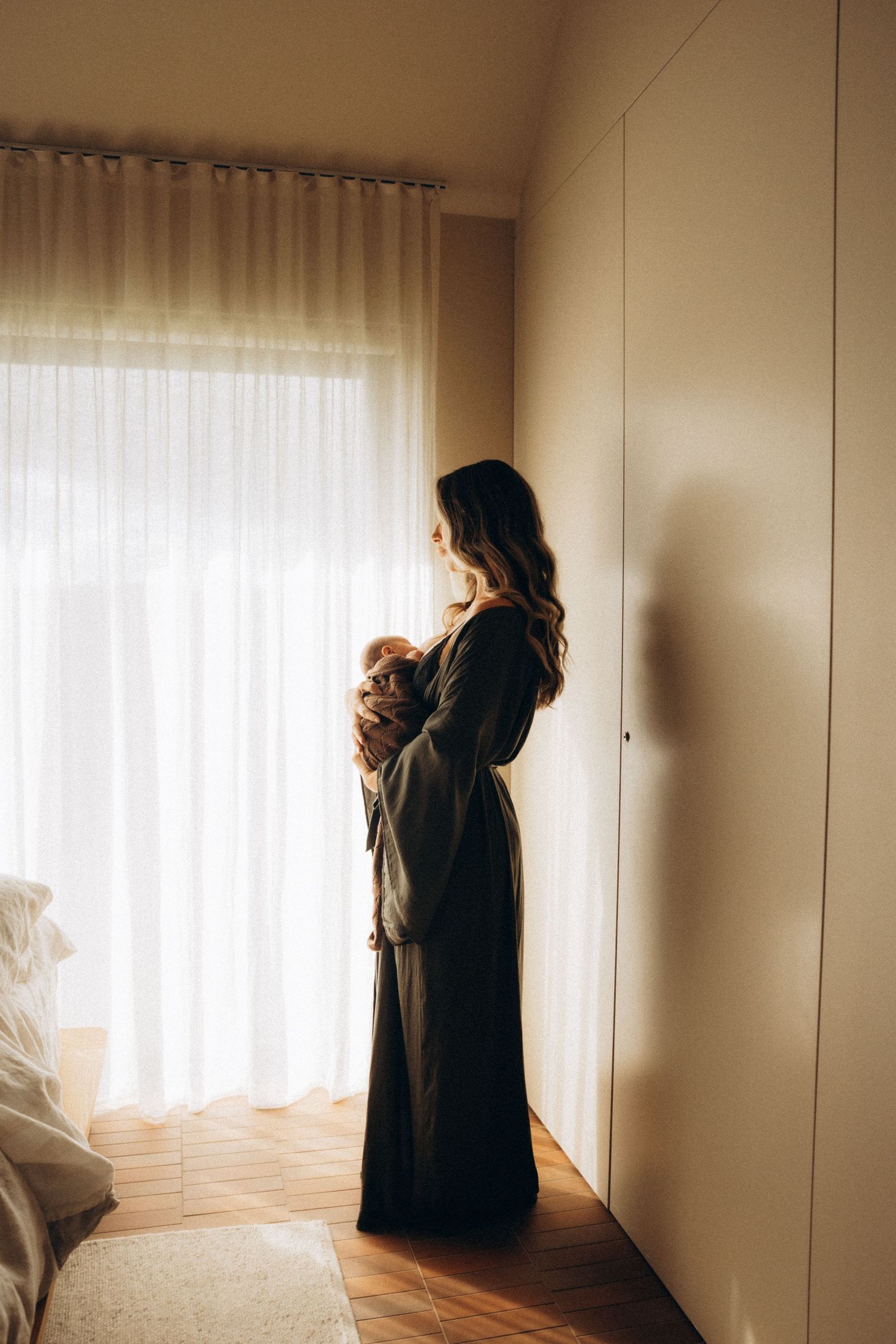 A couple embraces tenderly in front of a sunlit window with sheer curtains.