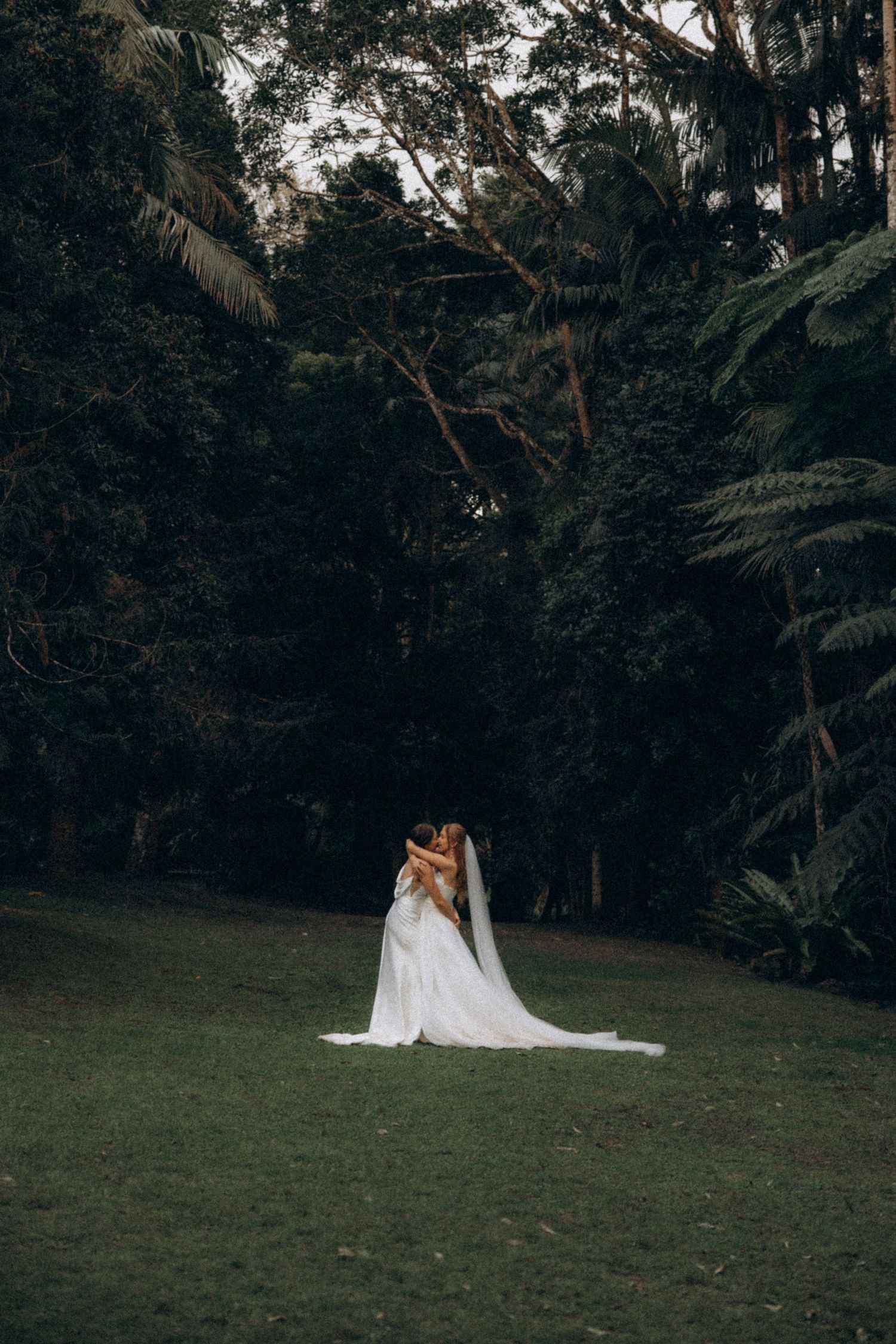 Two brides embrace in a lush green forest clearing, white gown and veil trailing.