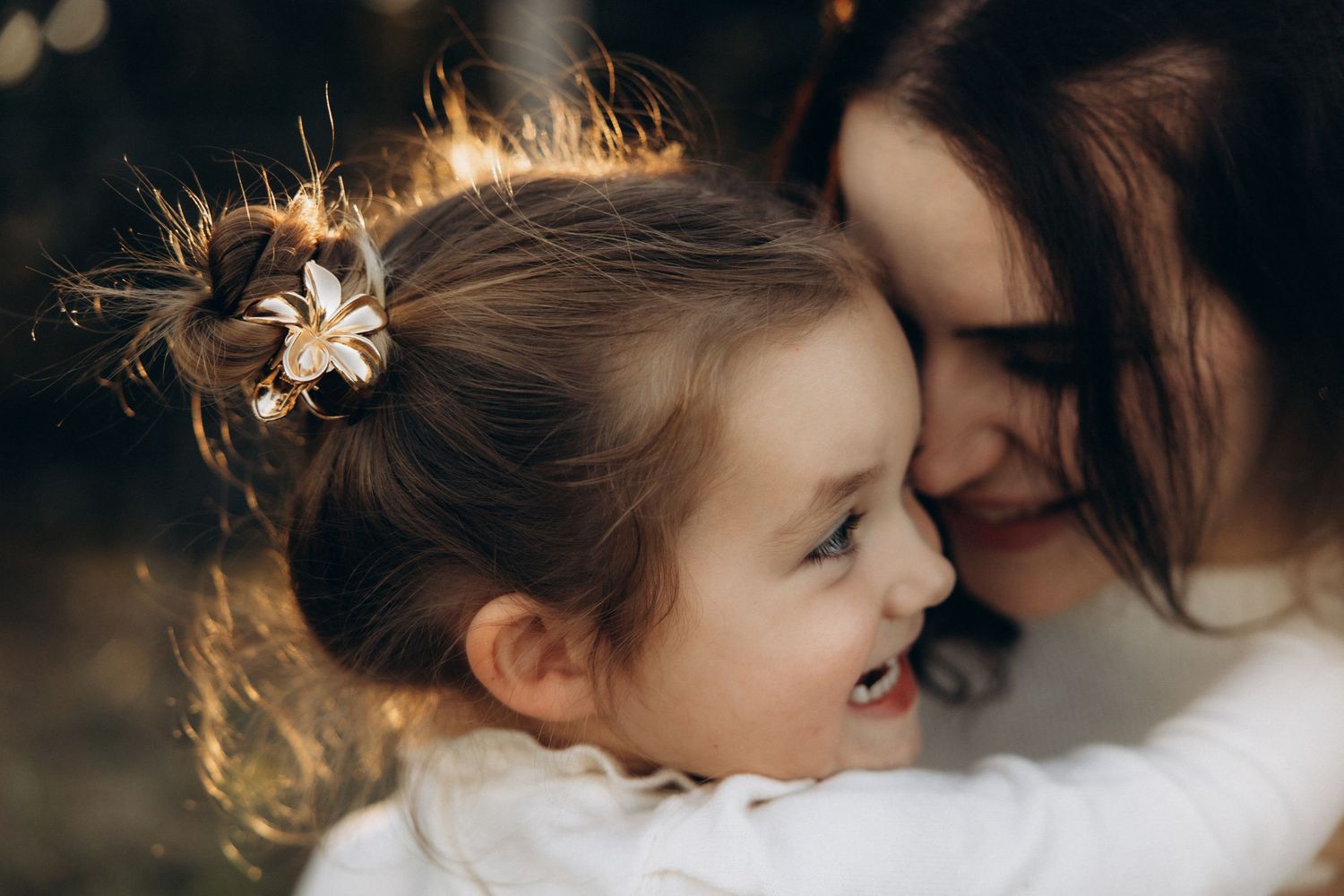 A mother and young daughter share a joyful, tender moment together, with the smiling girl wearing a floral hair clip.