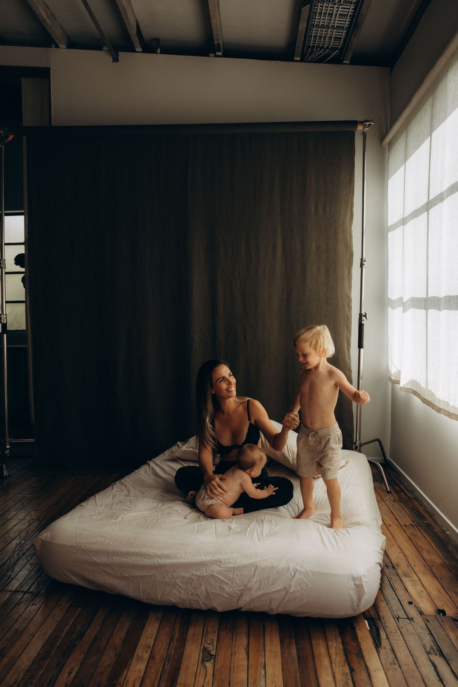 A mother plays with her two young children on a white mattress in a moody, loft-style room with wooden floors.