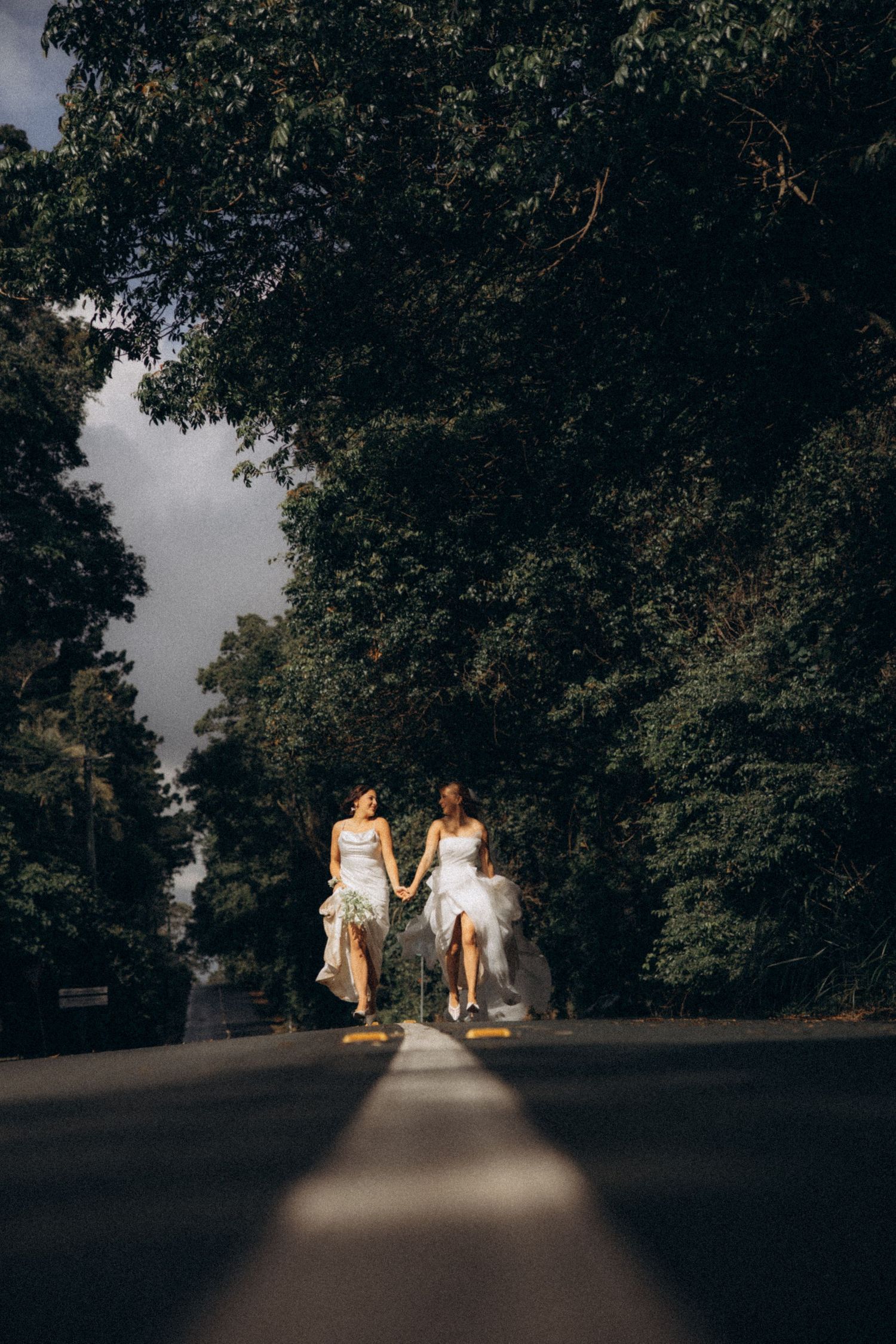 Two brides in white dresses hold hands running down a tree-lined road on their wedding day.