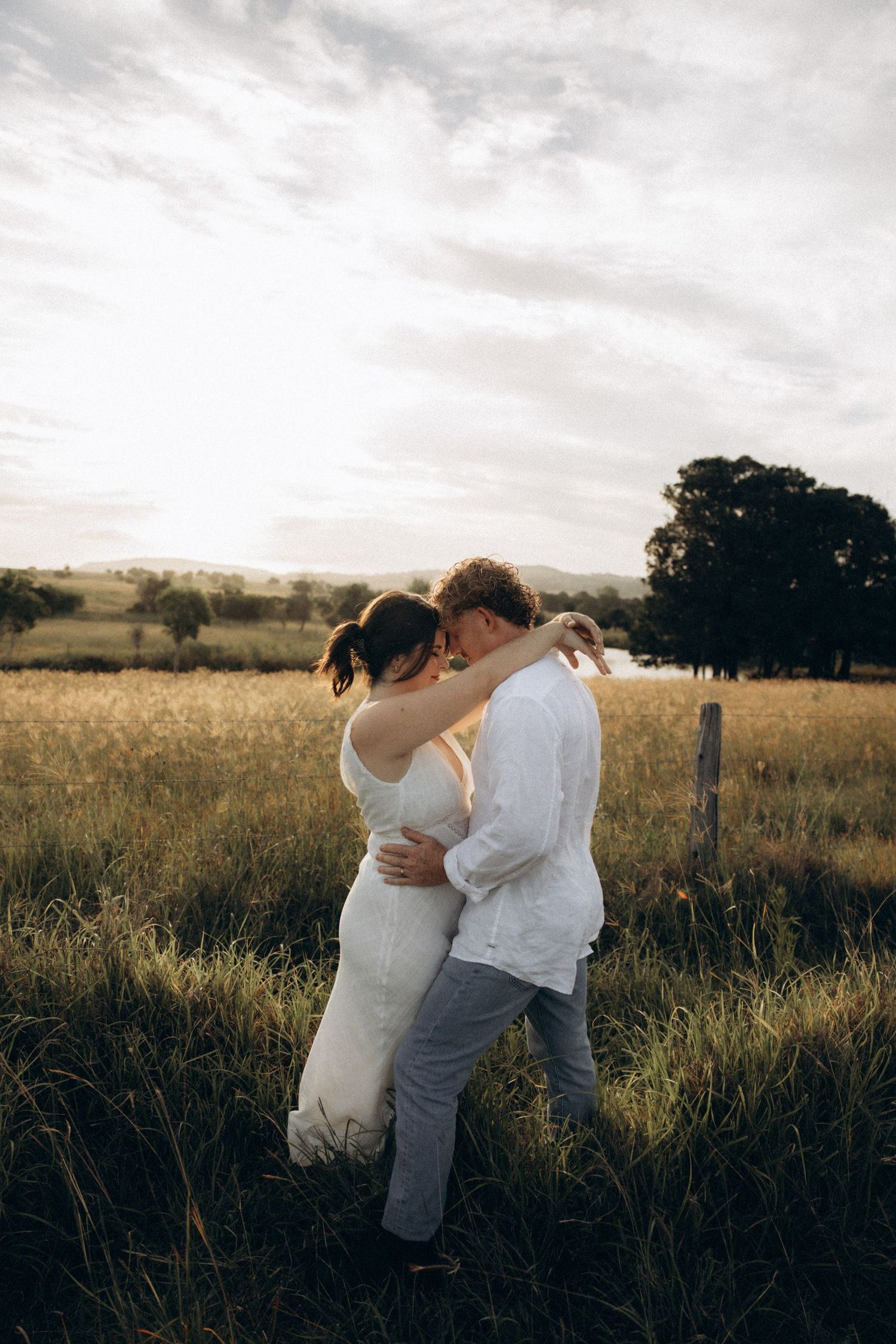 Couple in white clothing embraces in sunlit meadow with mountains in background.
