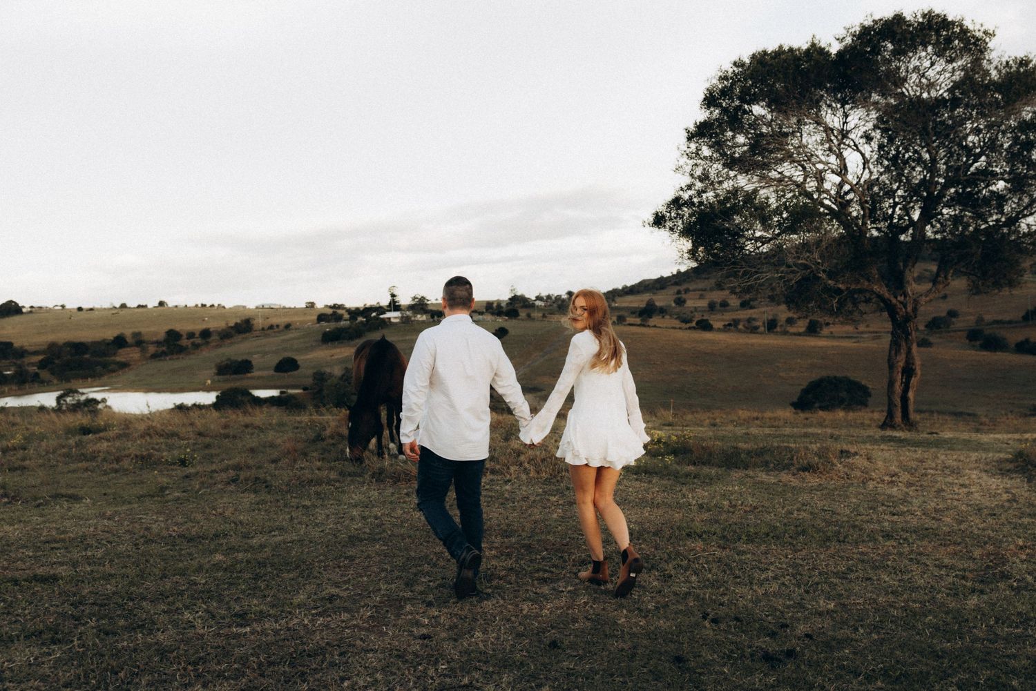 Couple holding hands walking through a rustic countryside field with trees and cattle in the background.