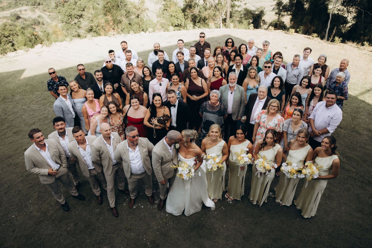 Large group photo of wedding party and guests on hillside in neutral attire.