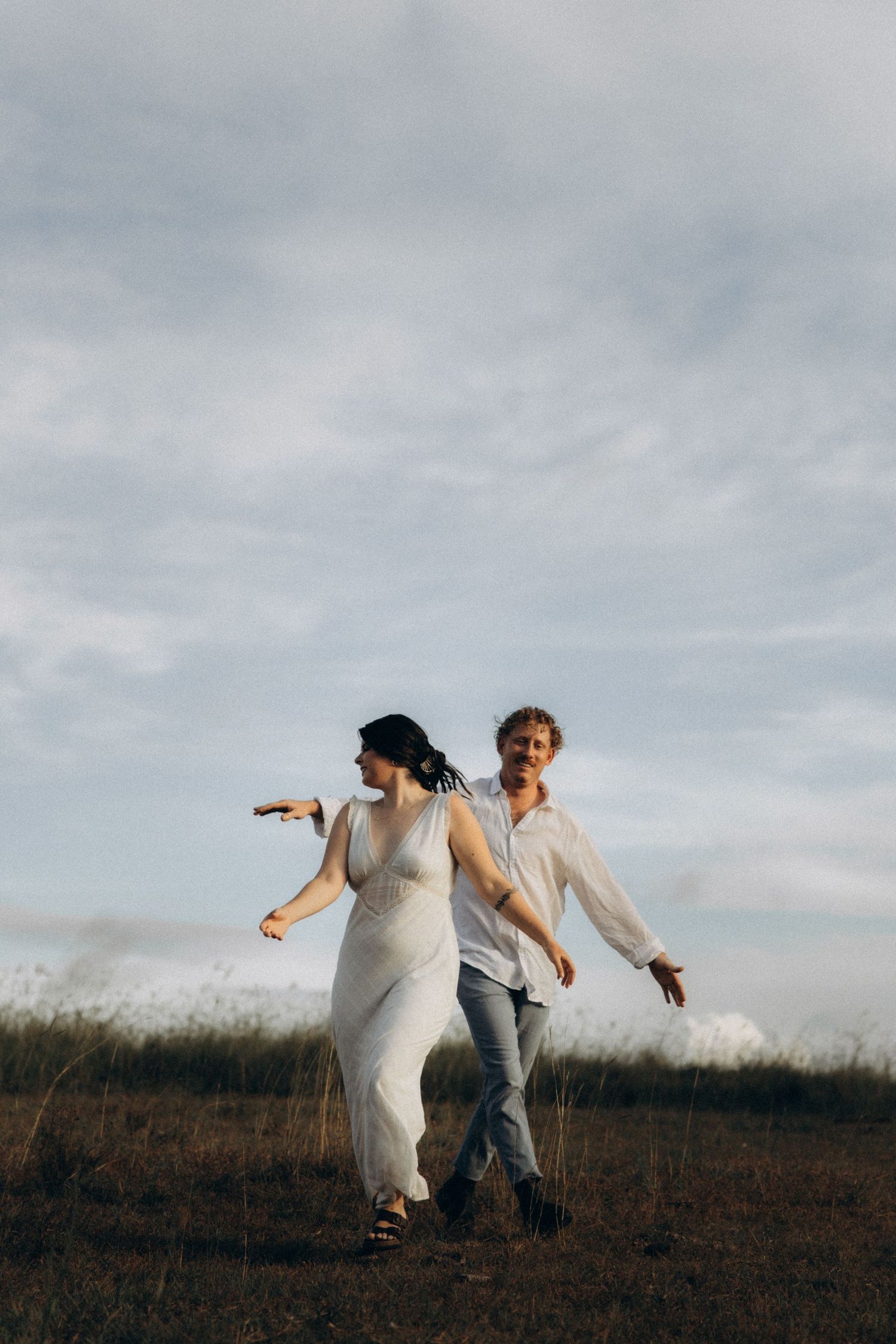 Two people run together through a rustic field under cloudy skies.