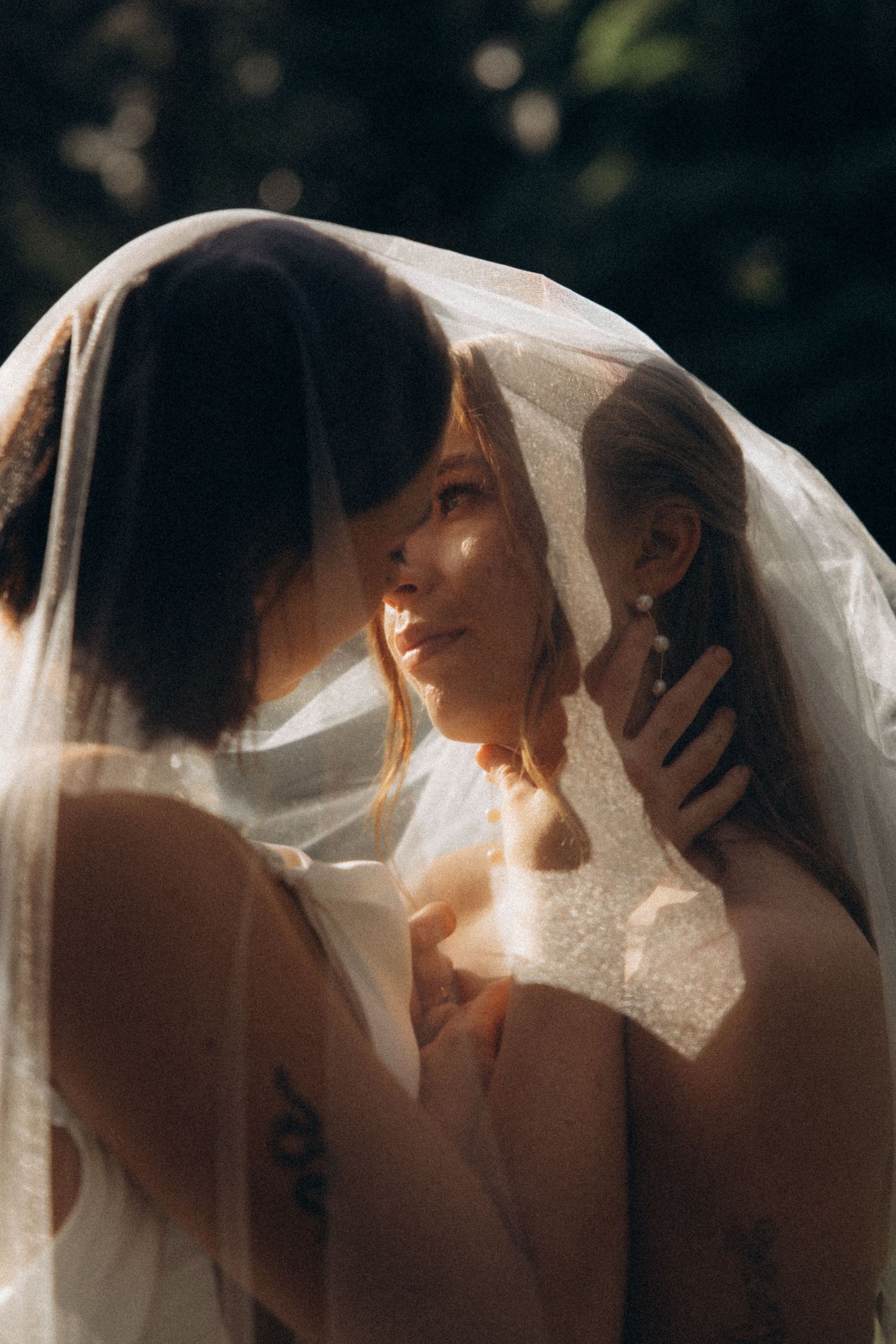 Two brides share a romantic moment under a flowing veil in dappled sunlight outdoors.