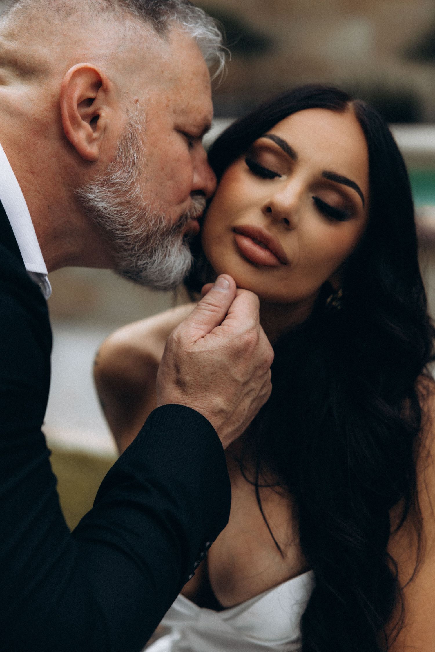 Elegant older man with gray beard kissing dark-haired bride's cheek during romantic wedding moment.