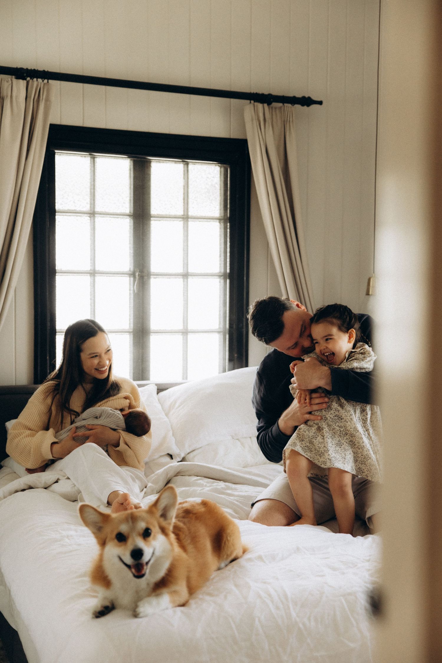 Family with newborn, toddler, and corgi dog relaxing together on a white bed near a bright window.