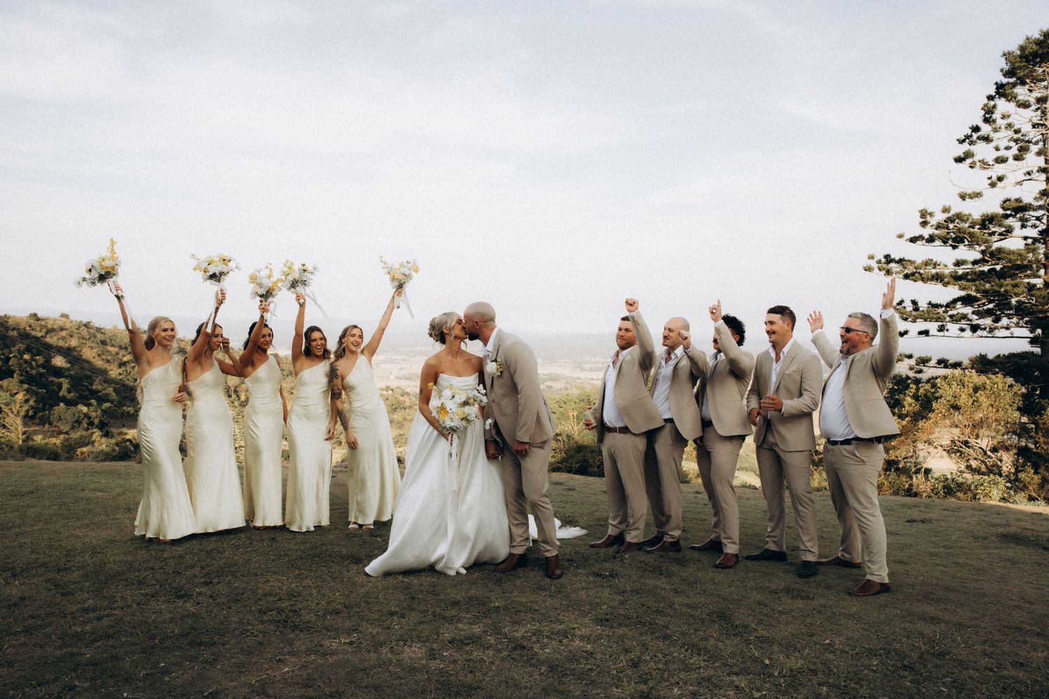 Wedding party in beige and white attire poses together on a scenic overlook.
