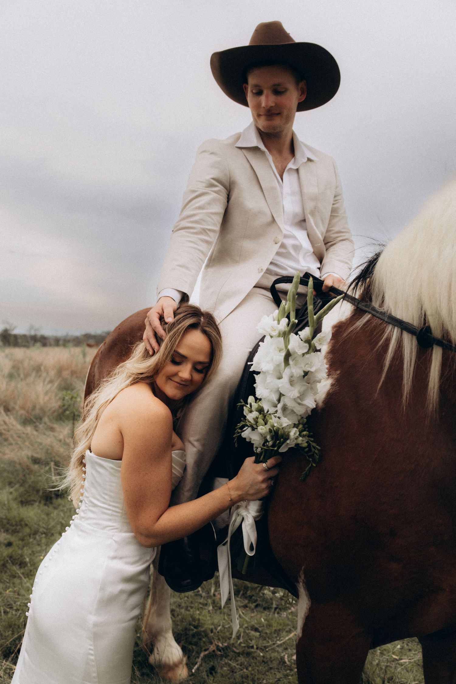 A couple in wedding attire poses with a horse in a rustic outdoor setting.