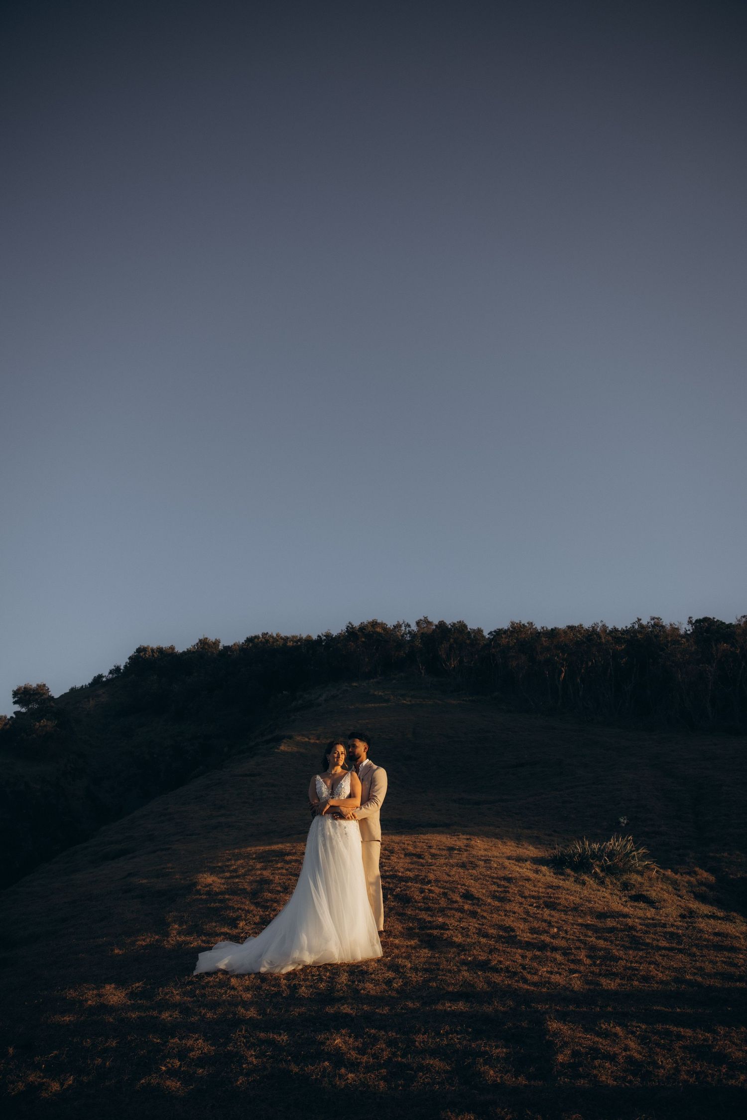 Two figures in wedding attire stand silhouetted against a dramatic sunset on a hilltop.
