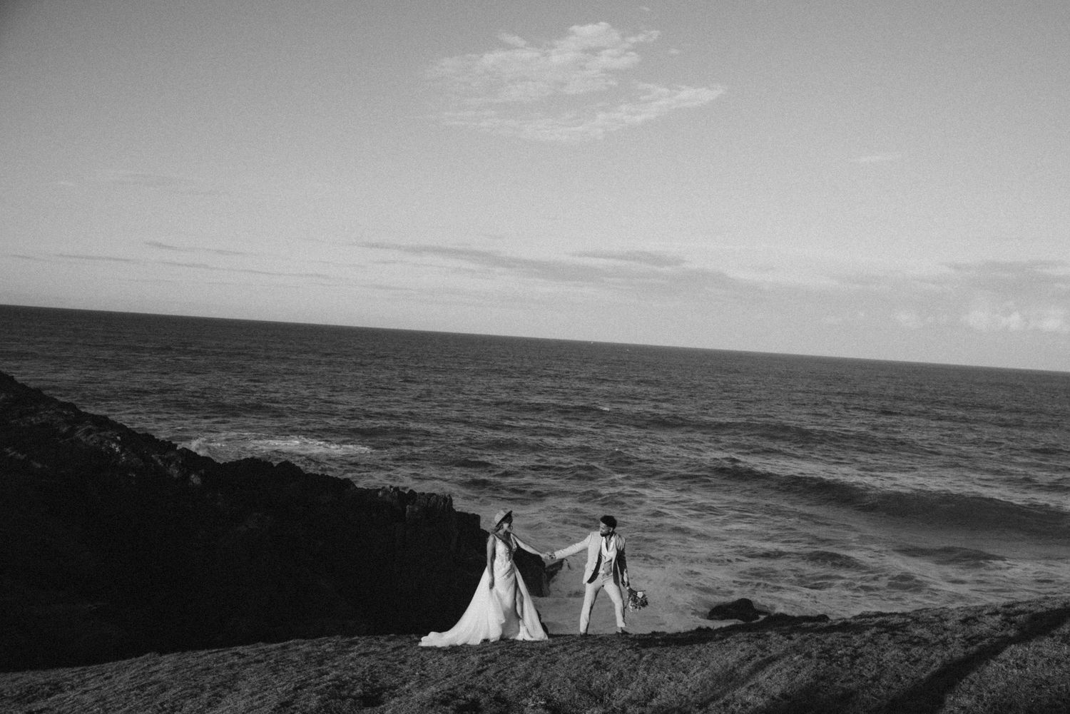 A dramatic black and white coastal landscape with wedding figures silhouetted against the ocean.