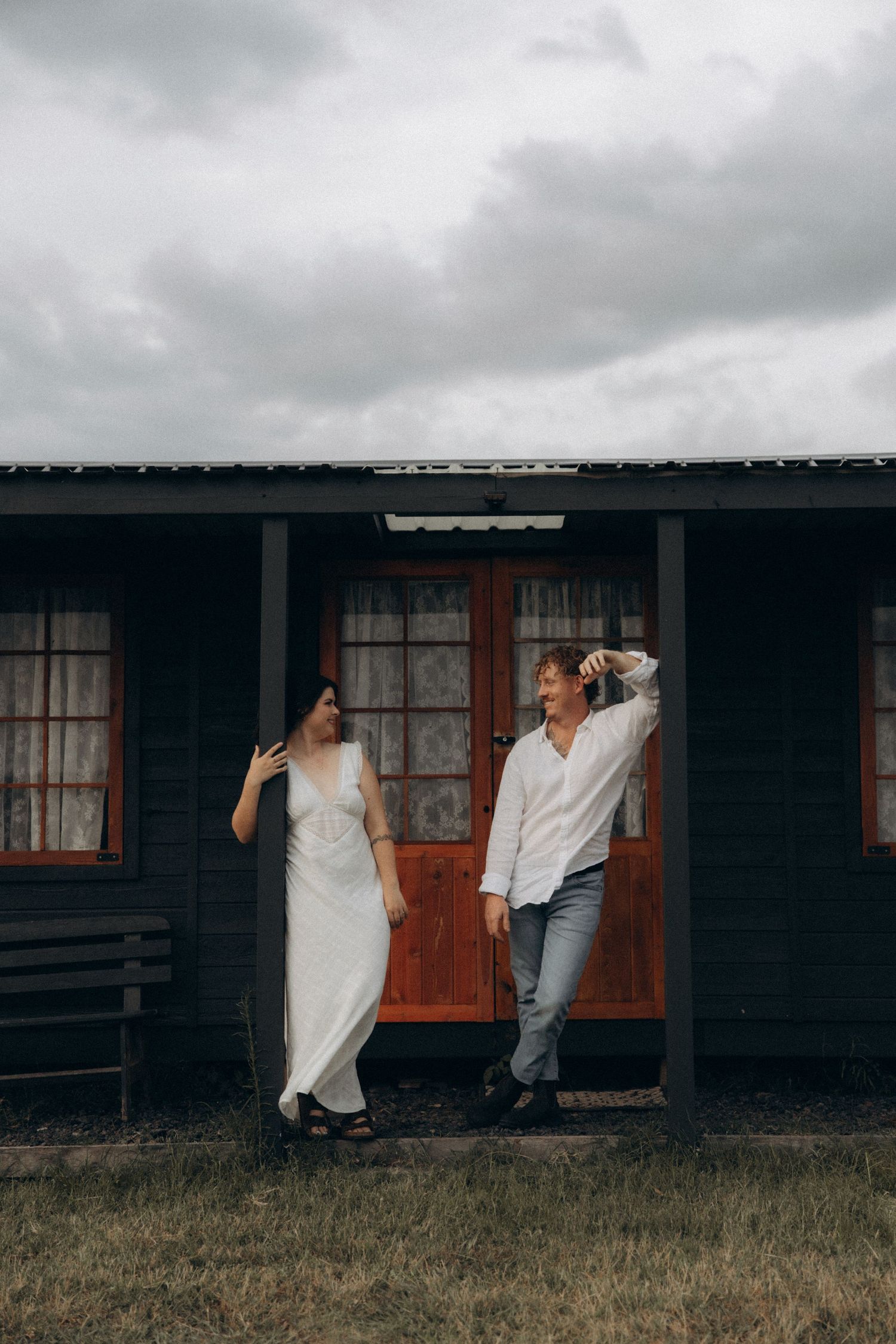 A couple poses casually outside a rustic cabin with wooden walls under cloudy skies.