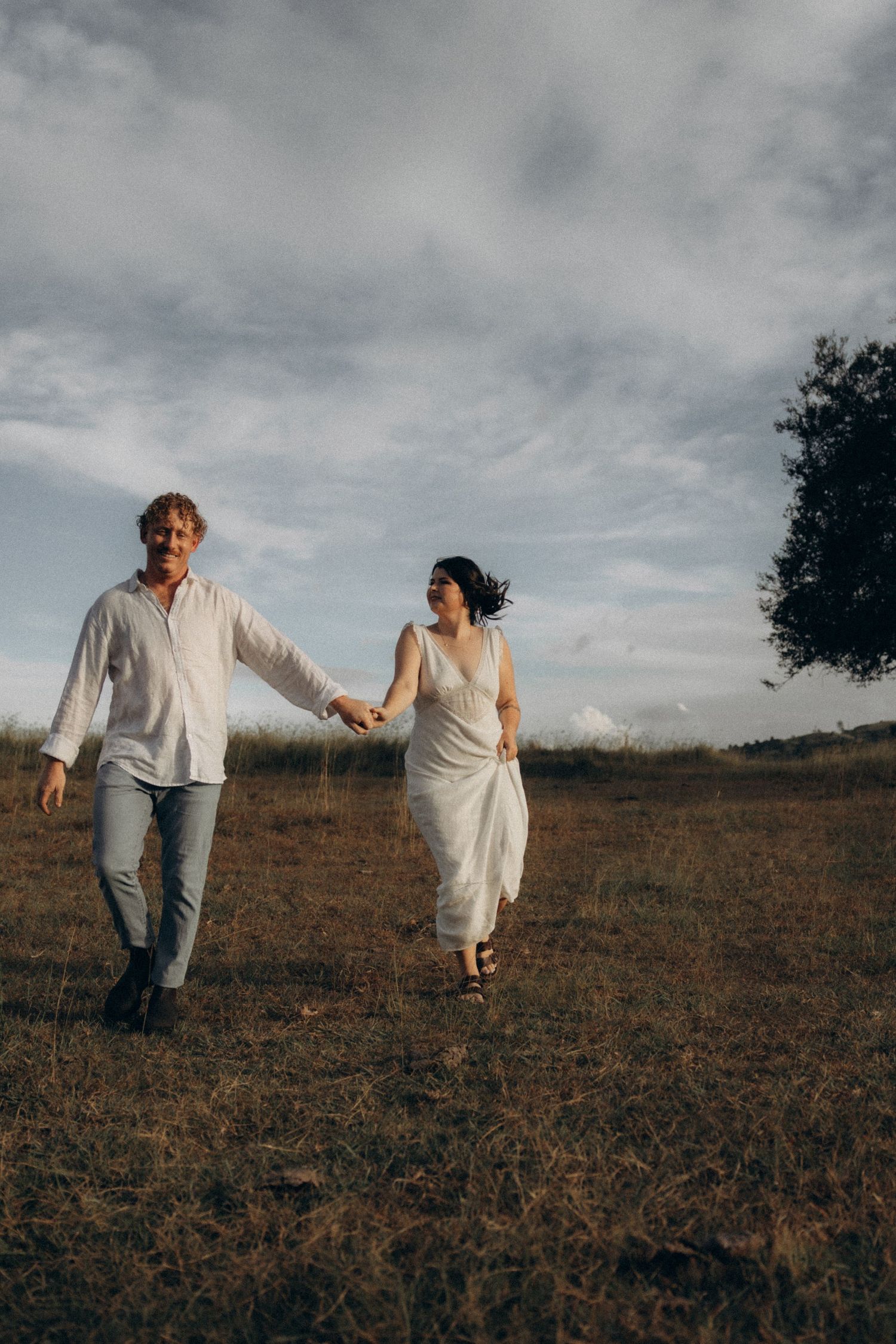 Two people hold hands while running through a golden field at dusk.