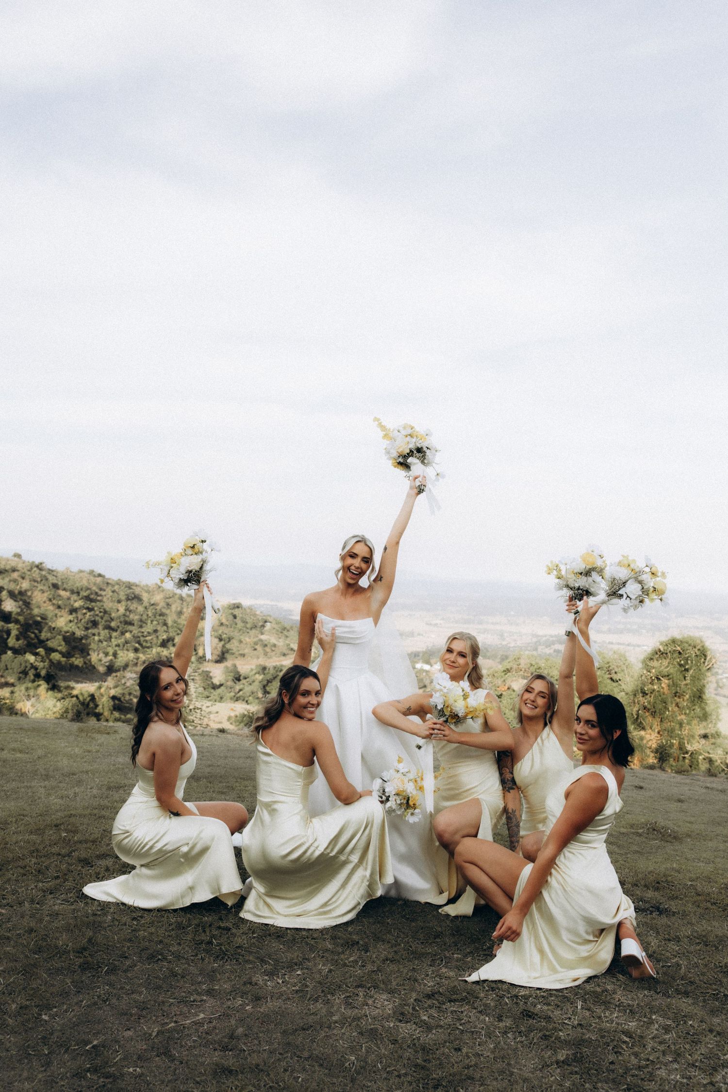 Bridesmaids in cream dresses pose playfully with bouquets on grassy hillside.