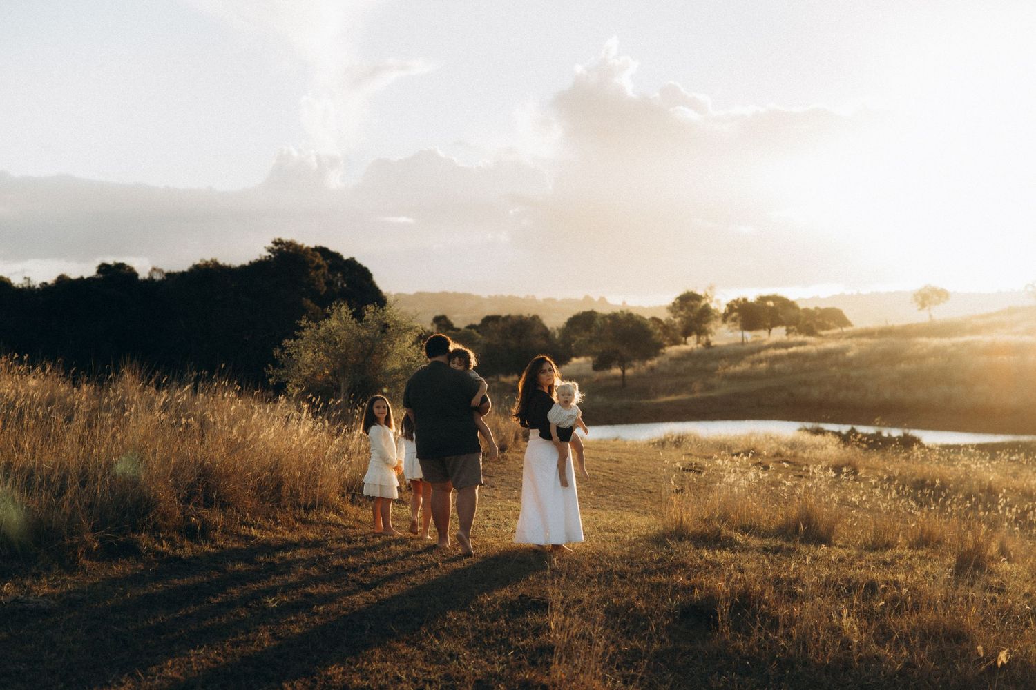 Family walking together at golden hour through a scenic countryside field with rolling hills and a pond in the background.