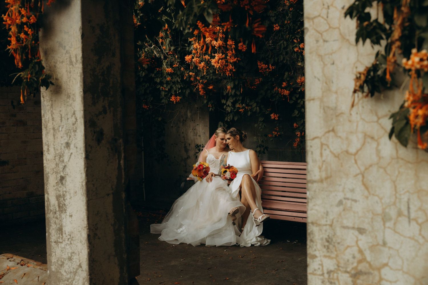 Bride and groom sitting on a pink bench surrounded by vibrant orange flowers at a romantic outdoor wedding venue.